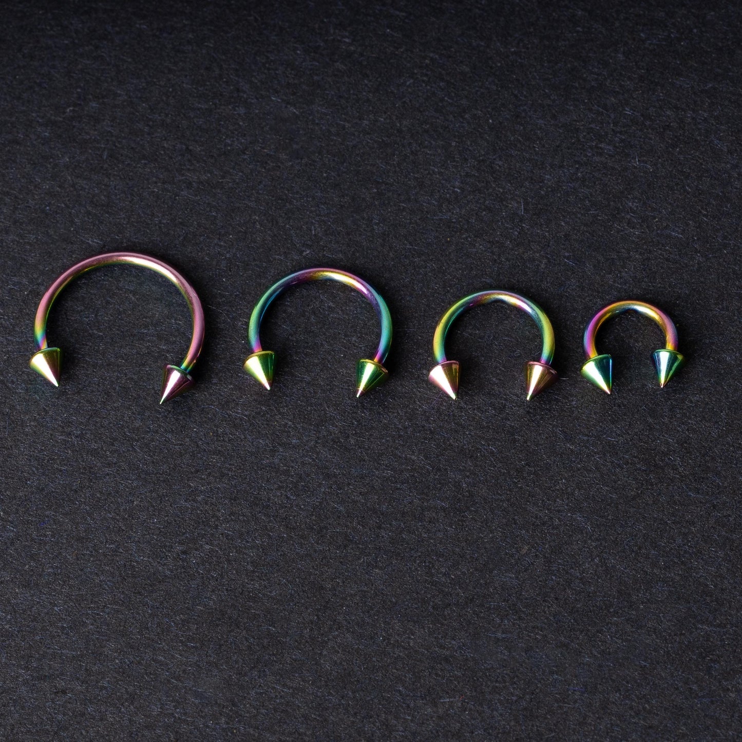 Titanium horseshoe hoops with spike ends, featuring a colorful rainbow finish, displayed on a dark textured background.