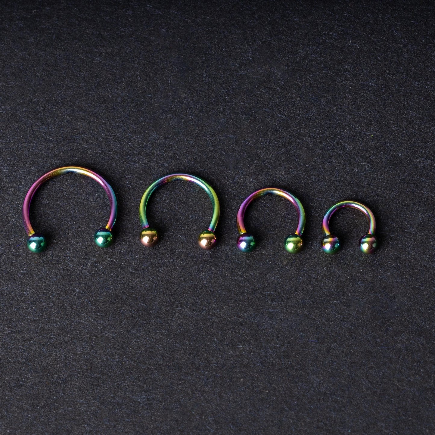 Close-up of four anodized rainbow horseshoe rings in varying sizes, with vibrant, multicolor finishes, displayed on a dark background.