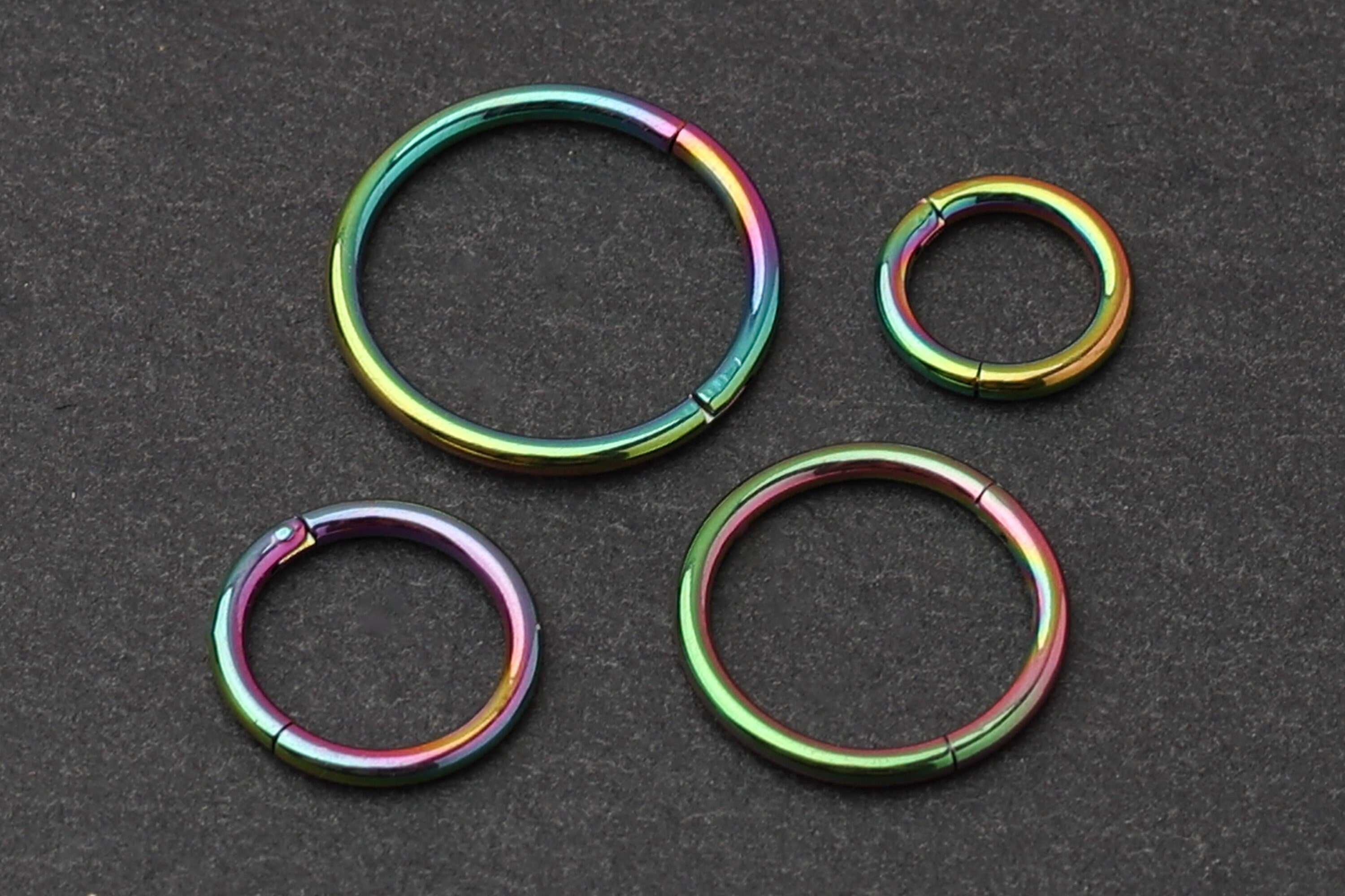 A collection of titanium seamless hoops in a vibrant rainbow finish, displayed on a dark textured background in varying sizes.
