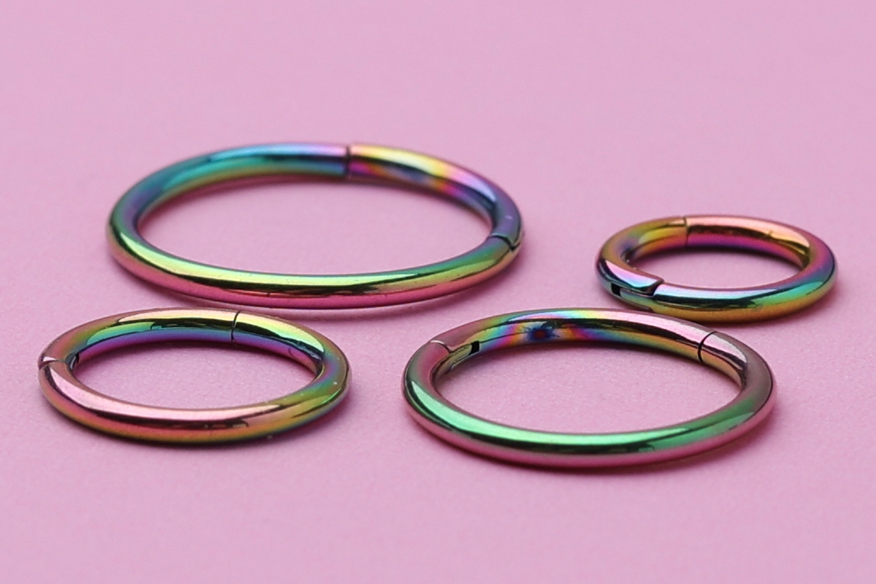 A collection of titanium seamless hoops in a vibrant rainbow finish, displayed on a Pink textured background in varying sizes.