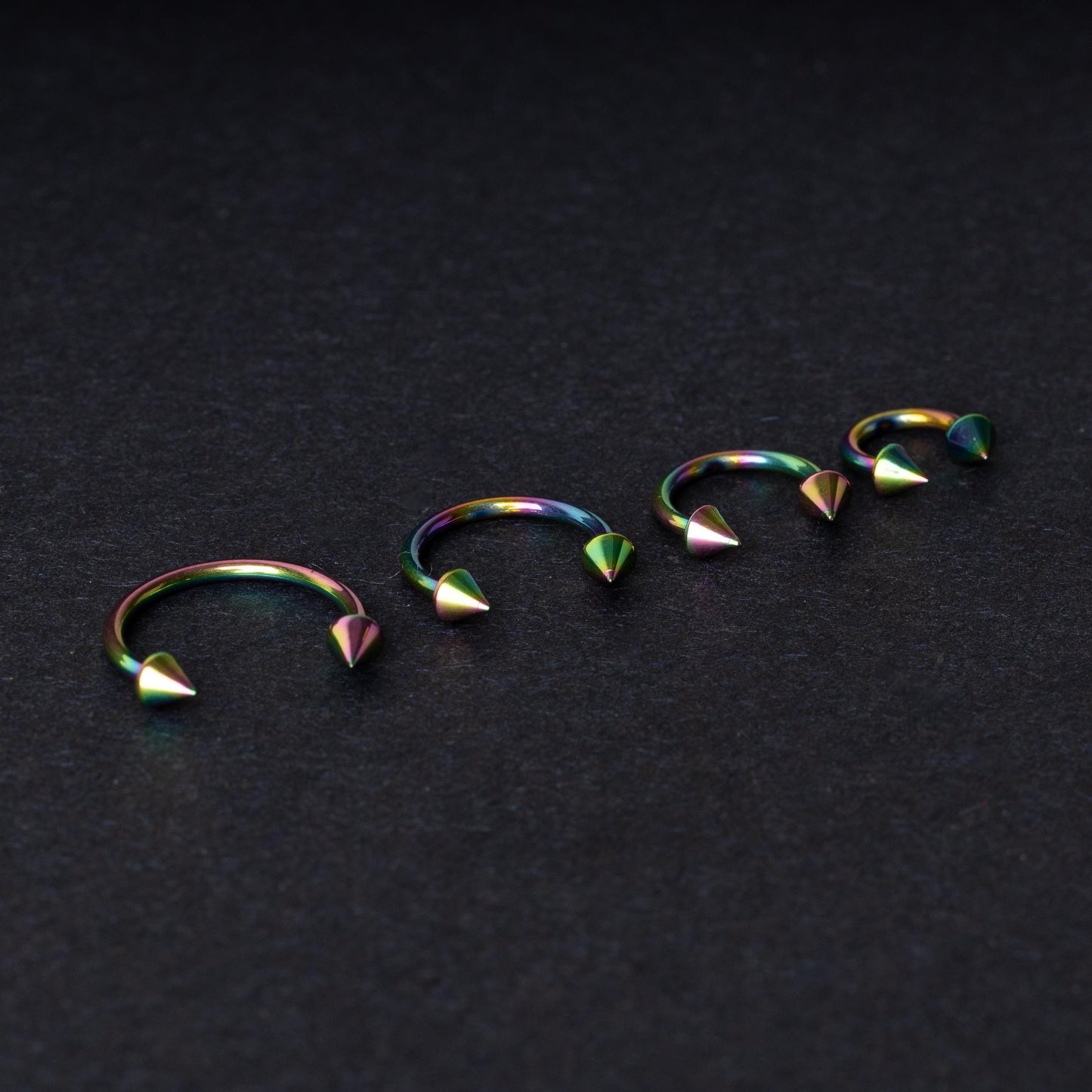 Titanium horseshoe hoops with spike ends, featuring a colorful rainbow finish, displayed on a dark textured background.