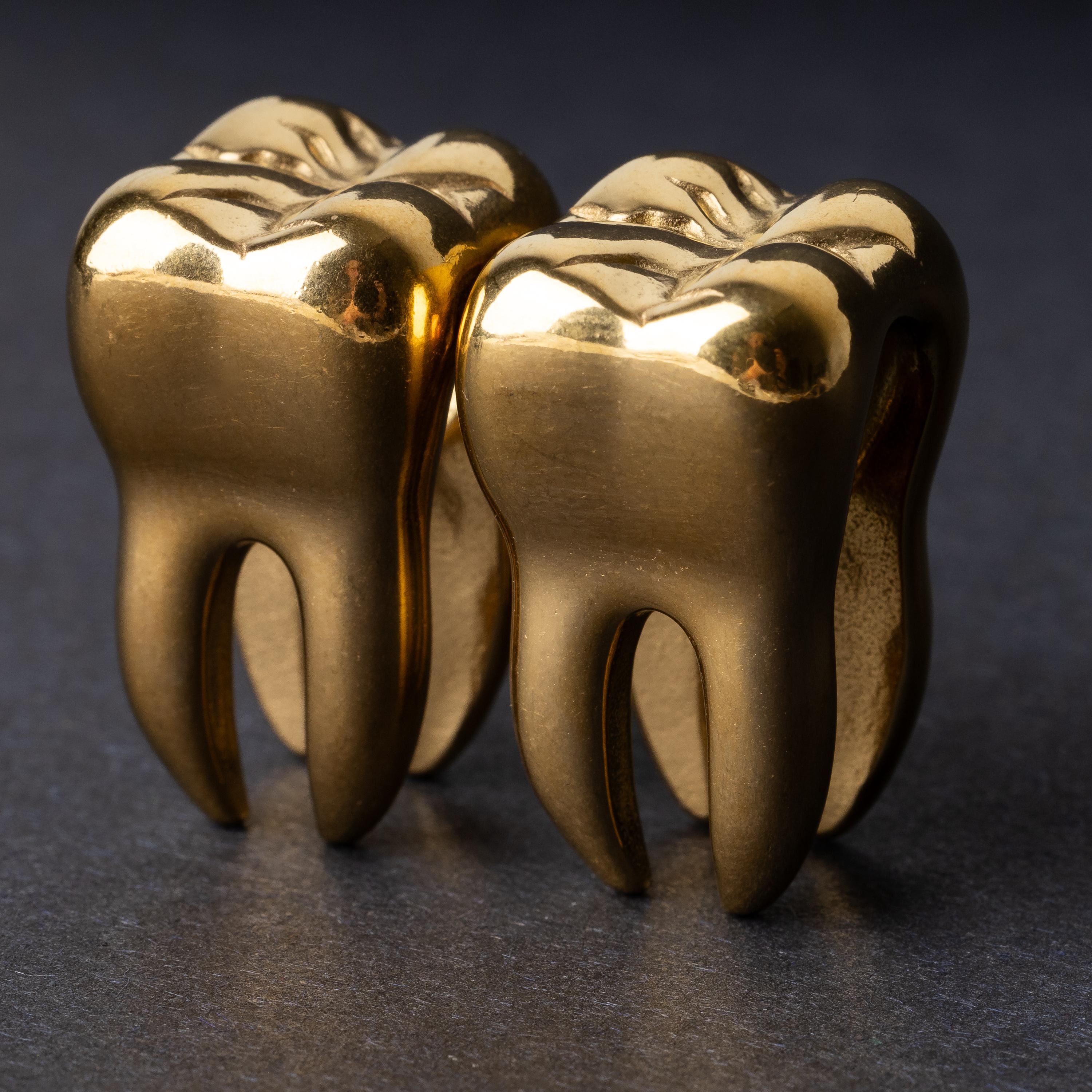 Close-up of gold tooth-shaped ear weights with polished detailing, displayed on a dark background.