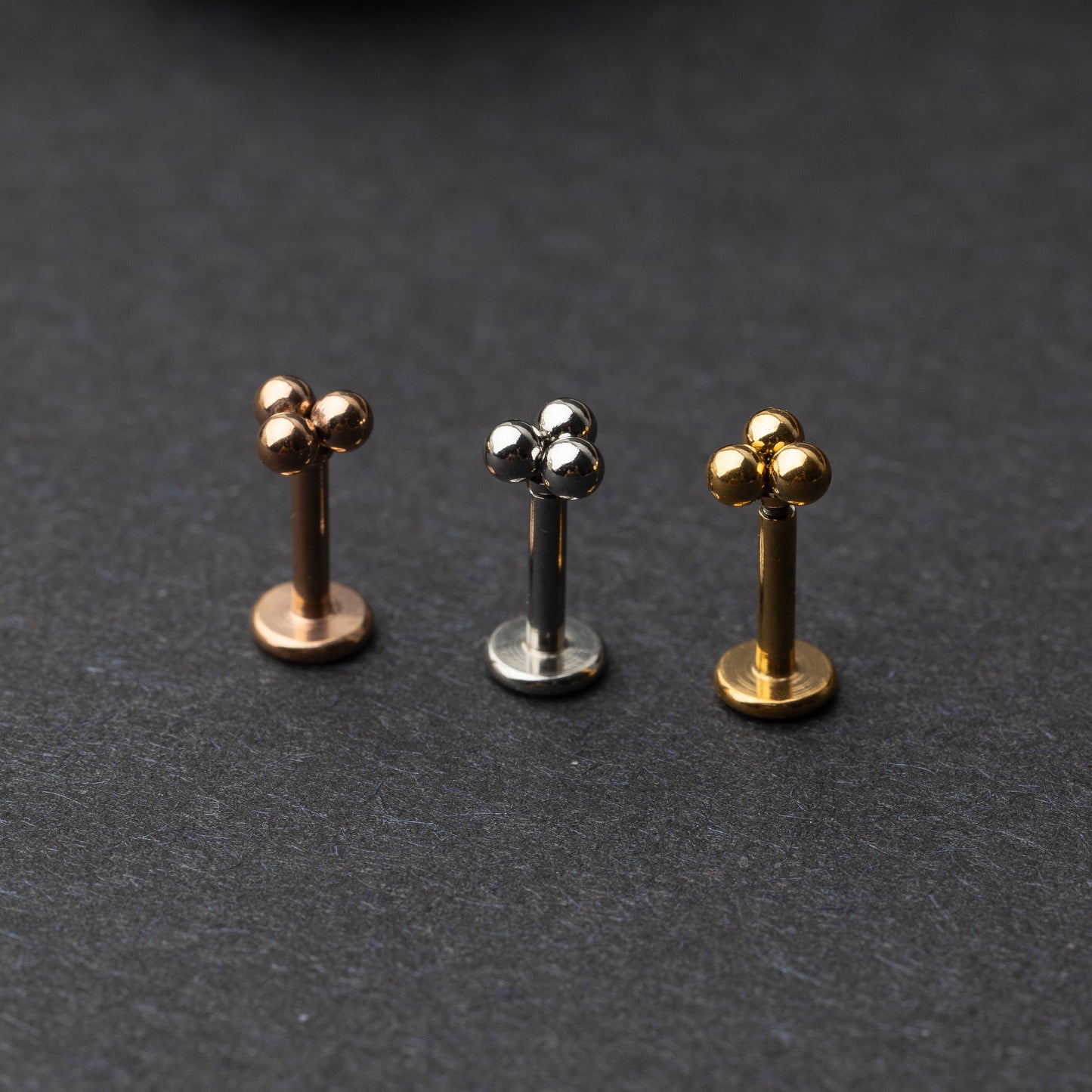 Close-up of three minimalist ball cluster labret studs in rose gold, silver, and gold finishes, displayed on a dark background.