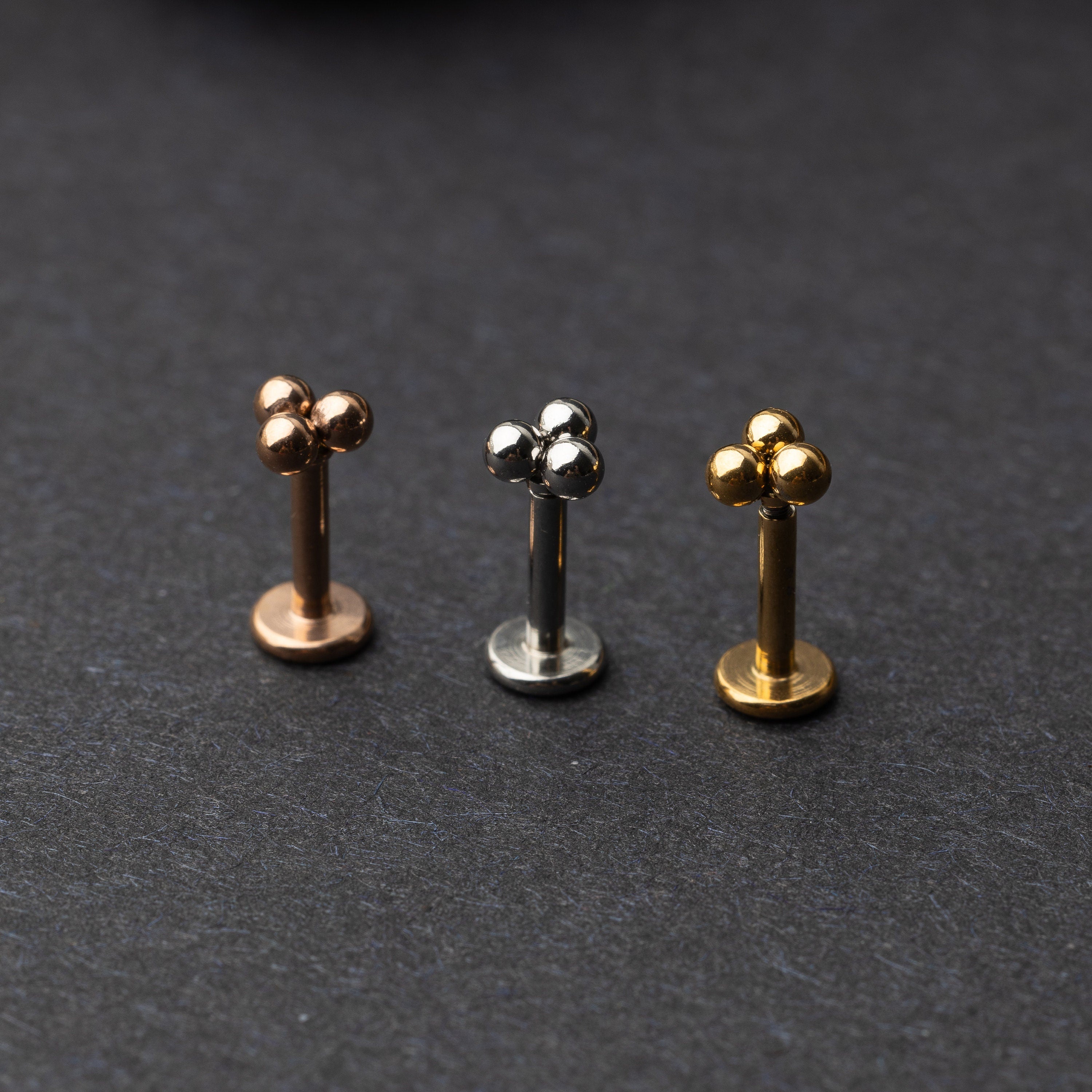 Close-up of three minimalist ball cluster labret studs in rose gold, silver, and gold finishes, displayed on a dark background.