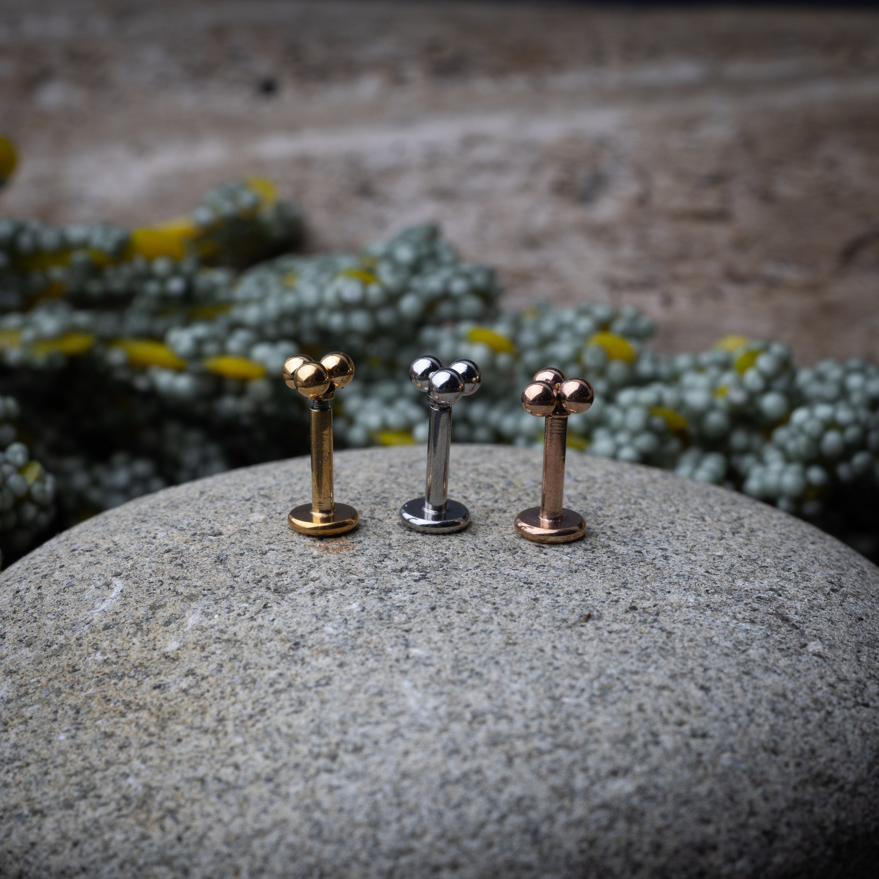 Close-up of three minimalist ball cluster labret studs in rose gold, silver, and gold finishes, displayed on a stone background.