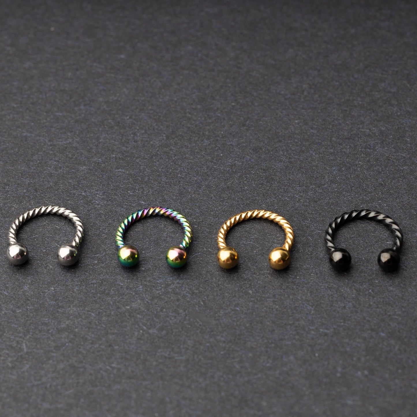 Twisted horseshoe barbells in silver, rainbow, gold, and black finishes displayed on a dark background, highlighting their bold and unique design.