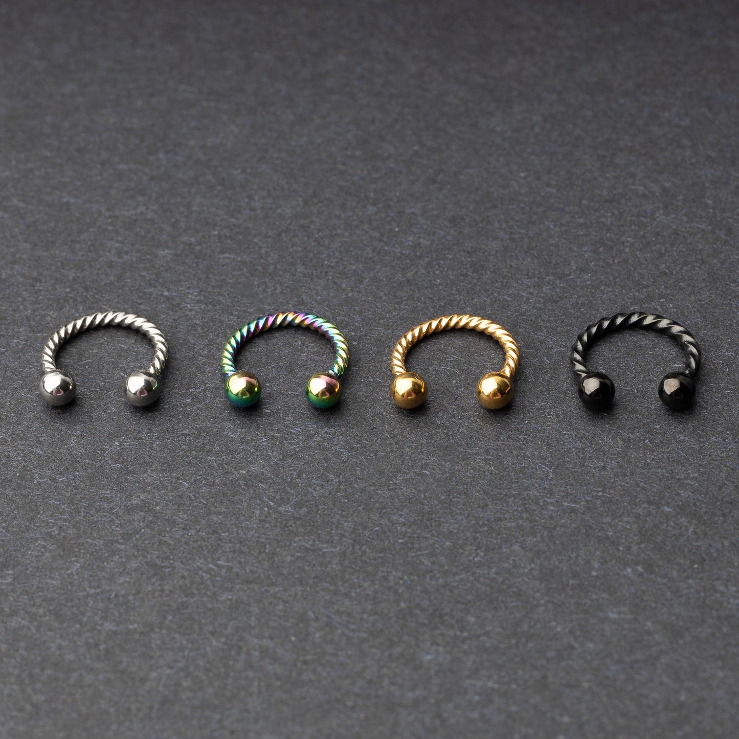 Twisted horseshoe barbells in silver, rainbow, gold, and black finishes displayed on a dark background, highlighting their bold and unique design.