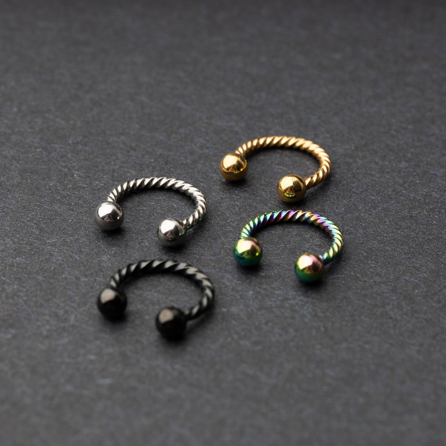 Twisted horseshoe barbells in silver, rainbow, gold, and black finishes displayed on a dark background, highlighting their bold and unique design.