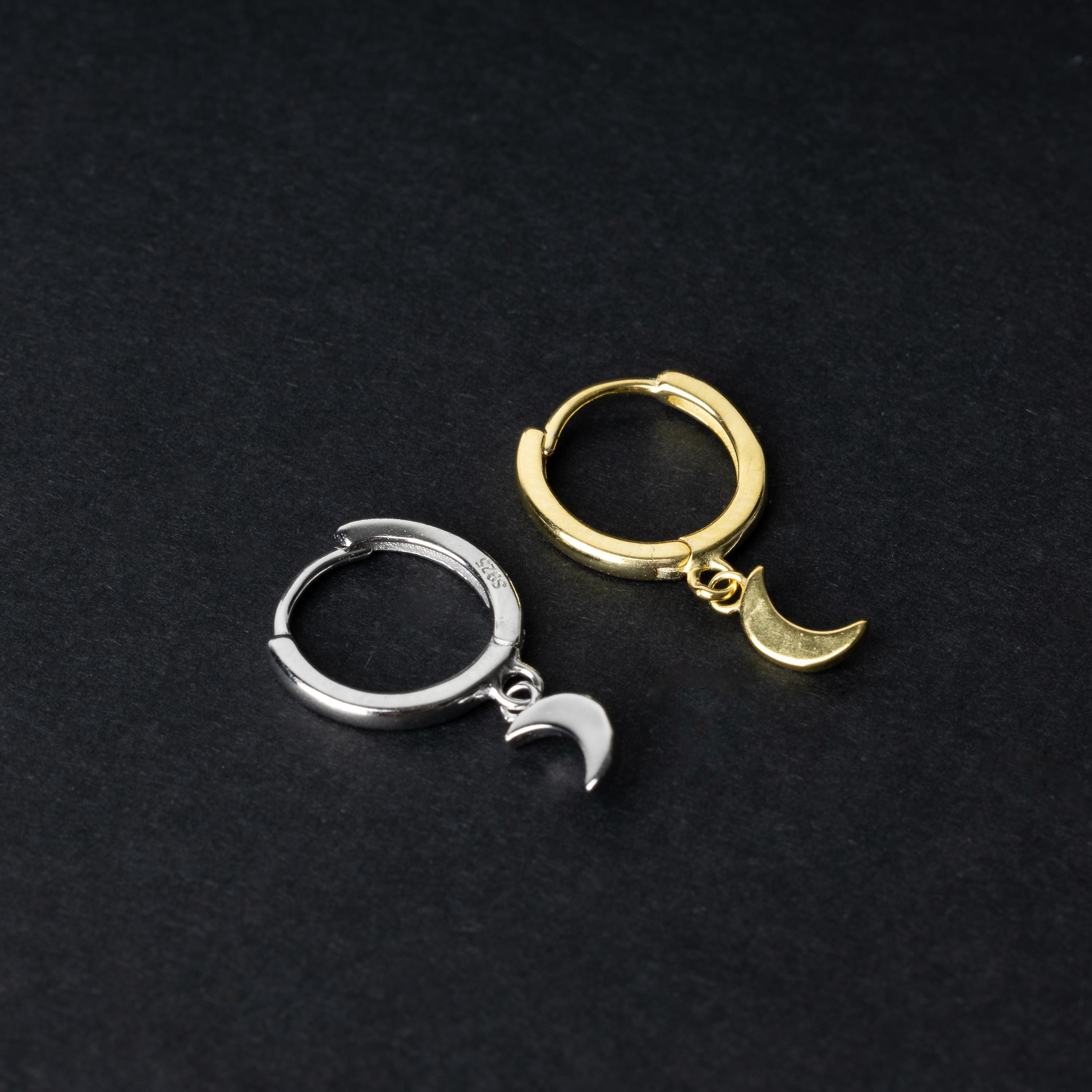 Two crescent moon huggie hoop earrings in gold and silver, each featuring a dangling crescent charm, displayed on a dark background.