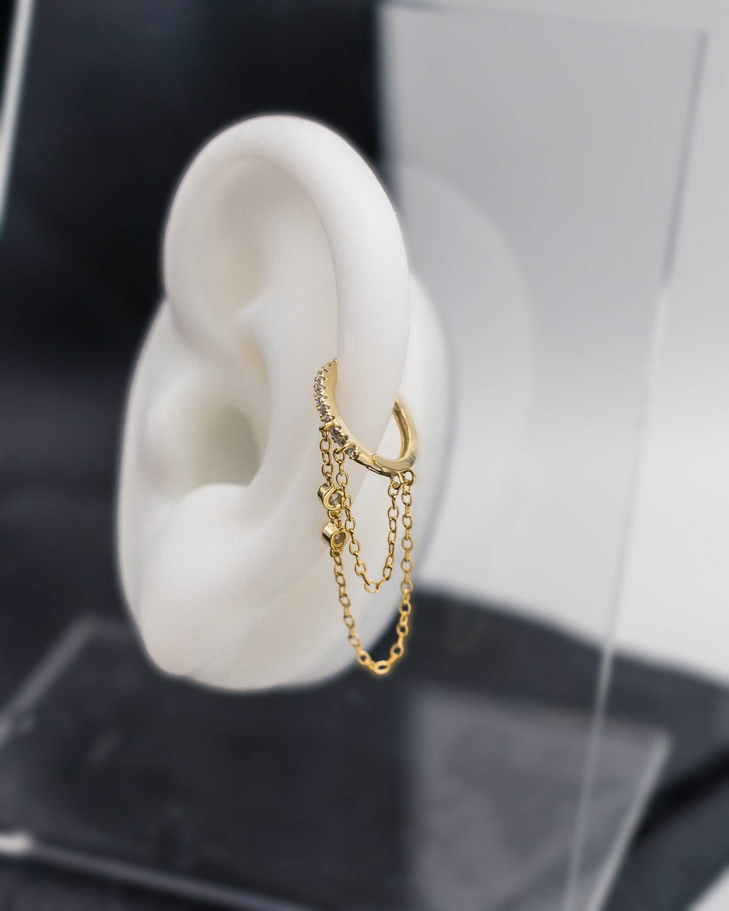 A gold septum clicker with a decorative design featuring dual chains and cubic zirconia gems, displayed on display ear