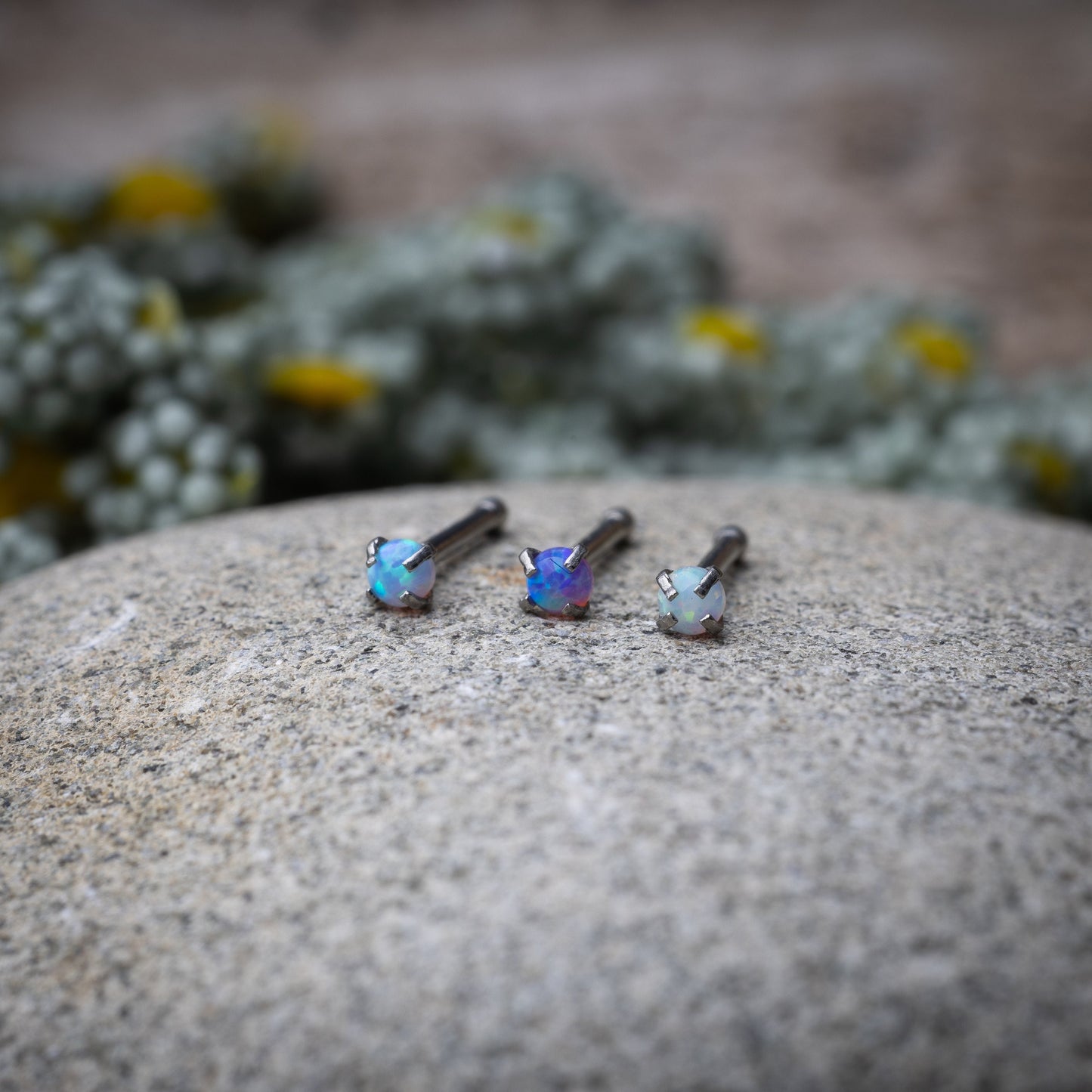 Three opalescent nose studs with prong settings, displayed on a stone background.