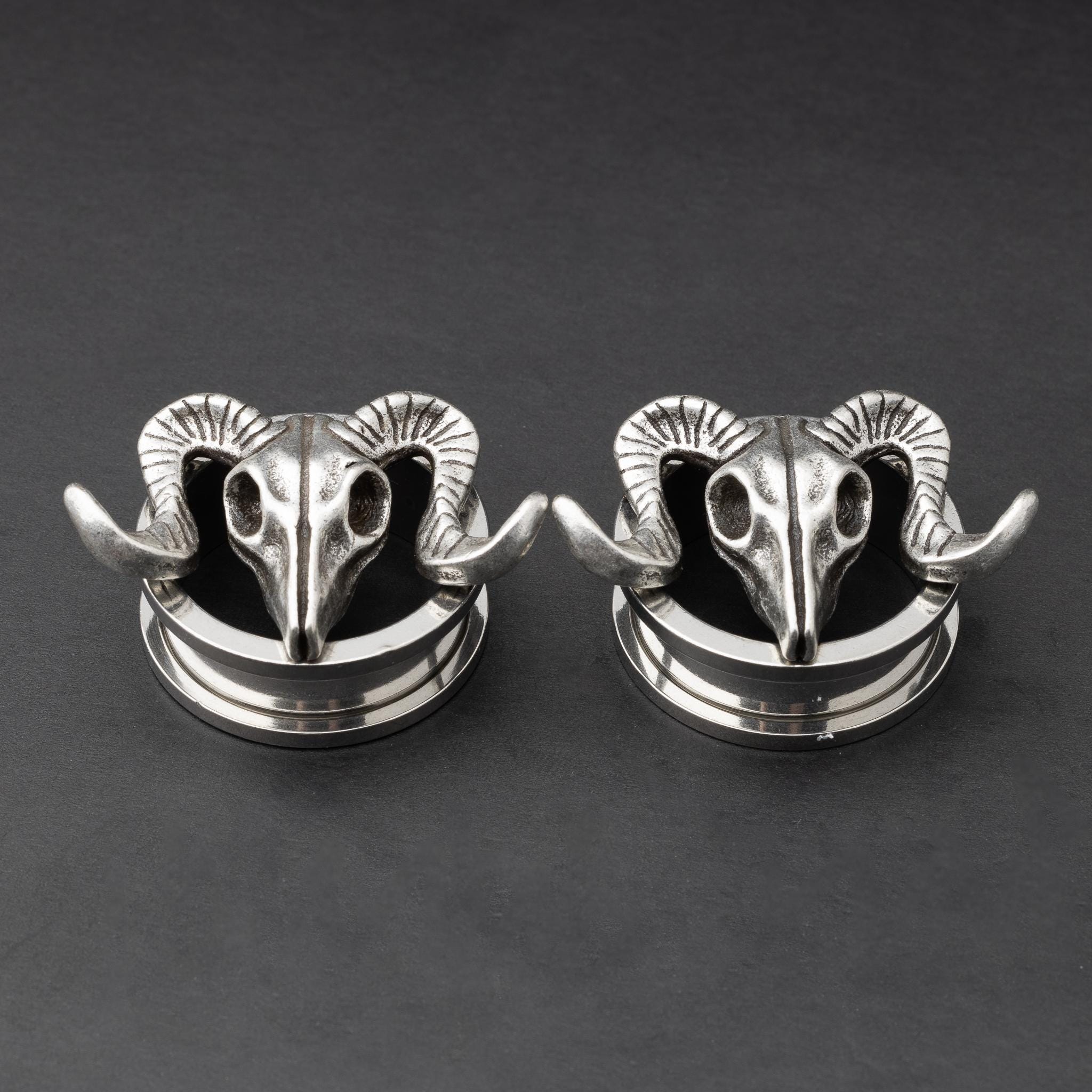 A pair of stainless steel saddle plugs featuring a 3D ram skull design, displayed on a dark, textured background.