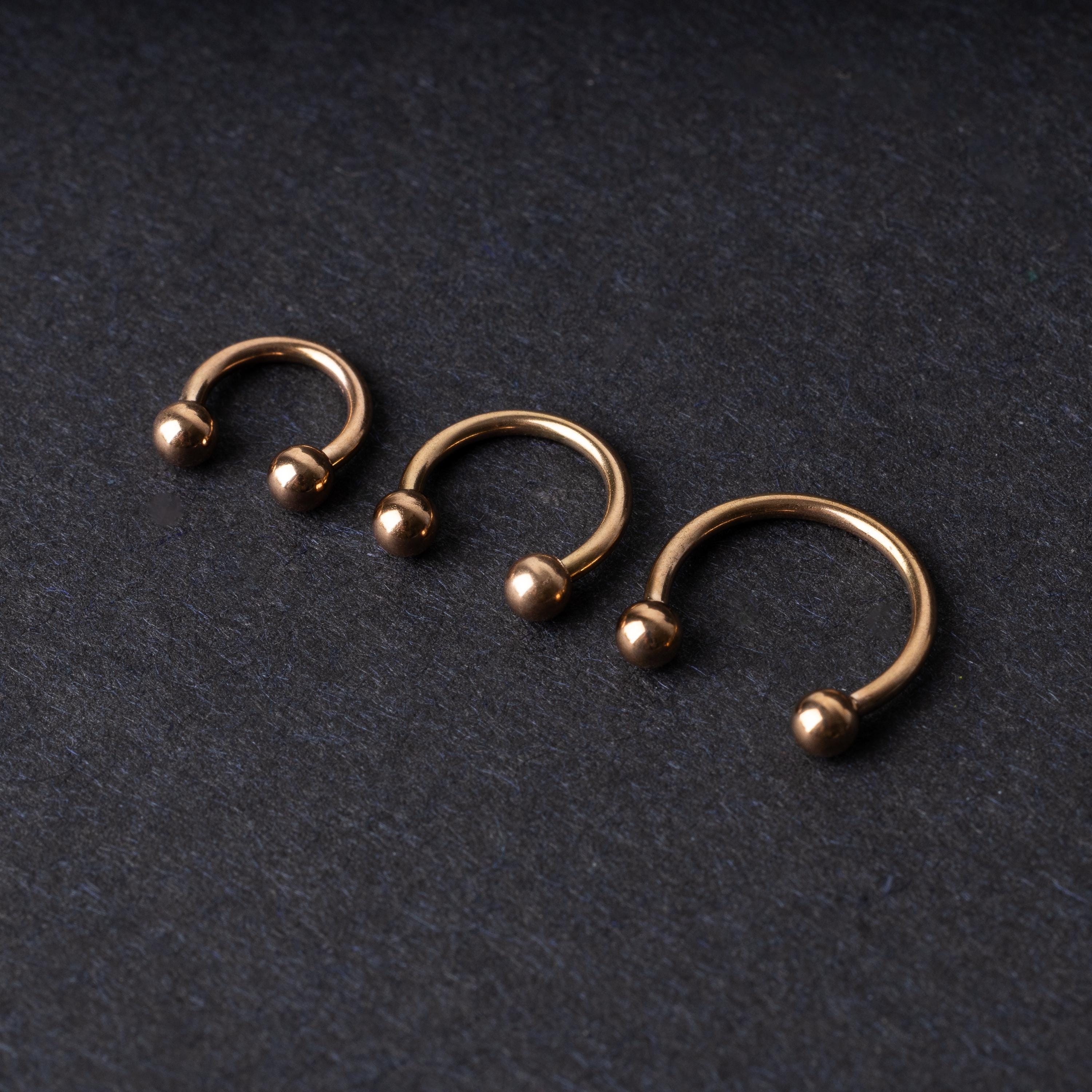 Three rose gold-tone stainless steel horseshoe hoops in varying sizes with screw-on ball ends, displayed against a dark background.