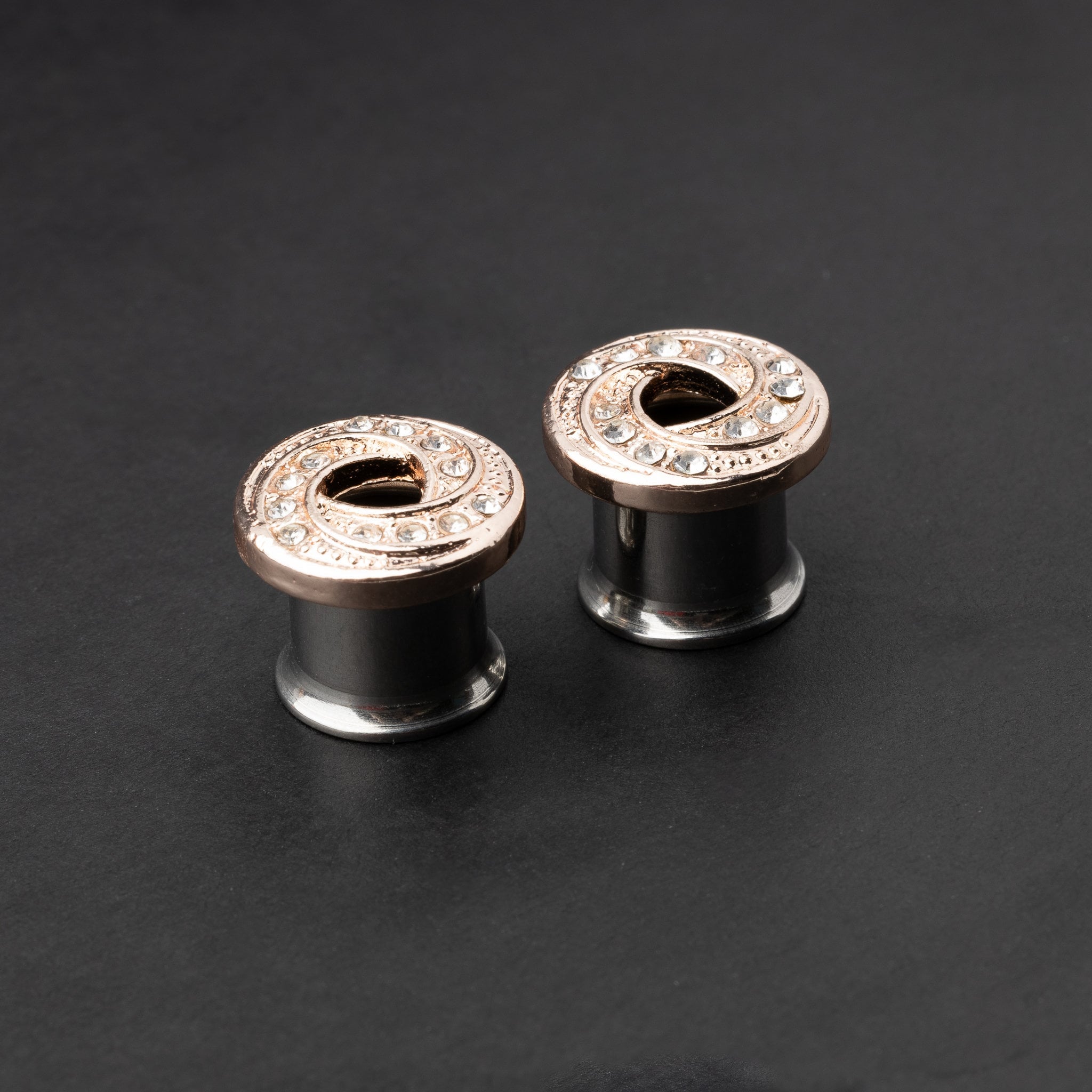 A pair of rose gold-tone stainless steel screw-fit tunnels with an infinity swirl design encrusted with clear crystals, displayed against a dark background.