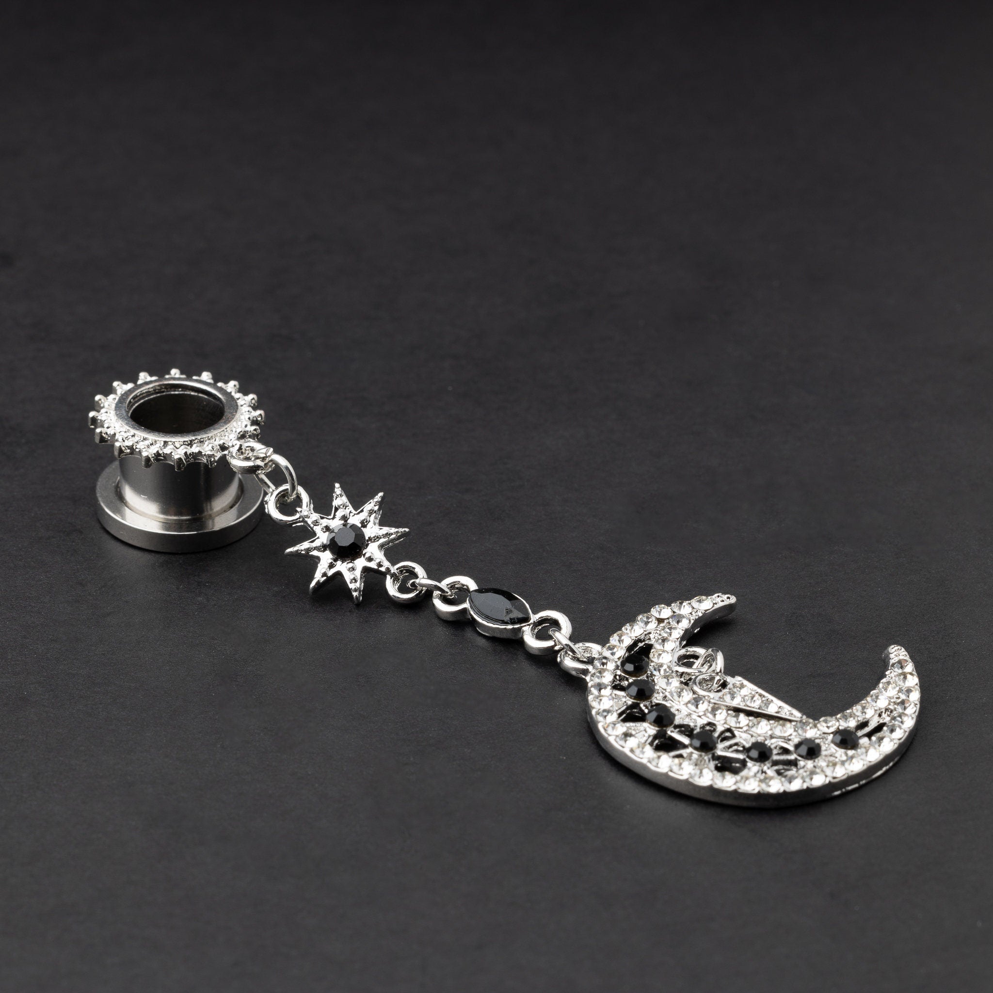 A pair of silver ear hangers for stretched ears with dangling crescent moon and star designs, adorned with black and clear rhinestones, displayed on a dark background.