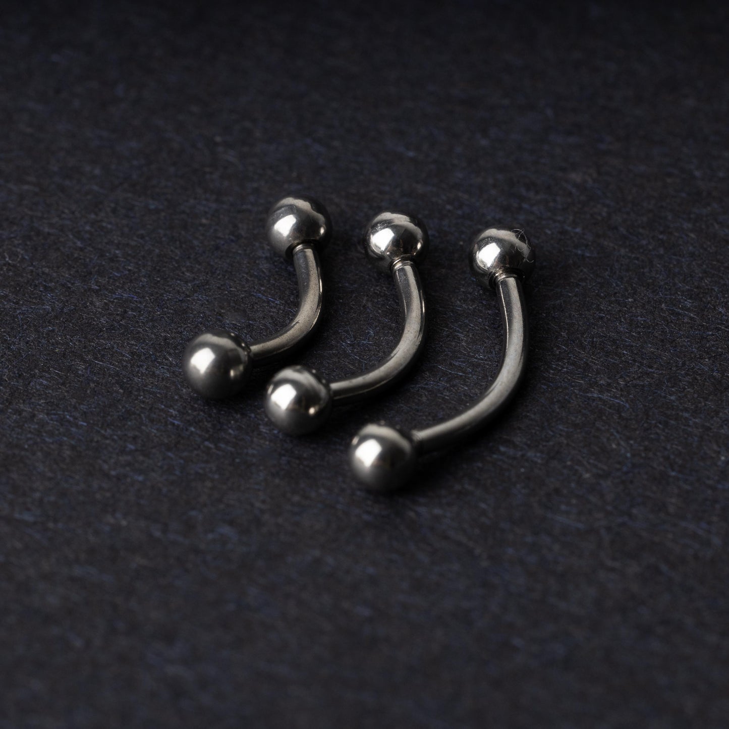 Three silver stainless steel curved barbells with polished ball ends, displayed on a dark textured background.
