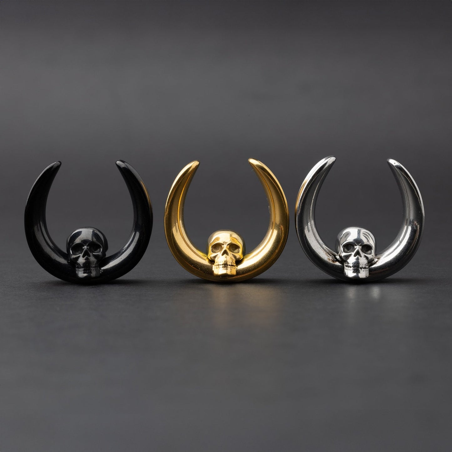 Three skull saddle tunnels in black, gold, and silver finishes displayed on a dark background, showcasing their bold and detailed skull design.