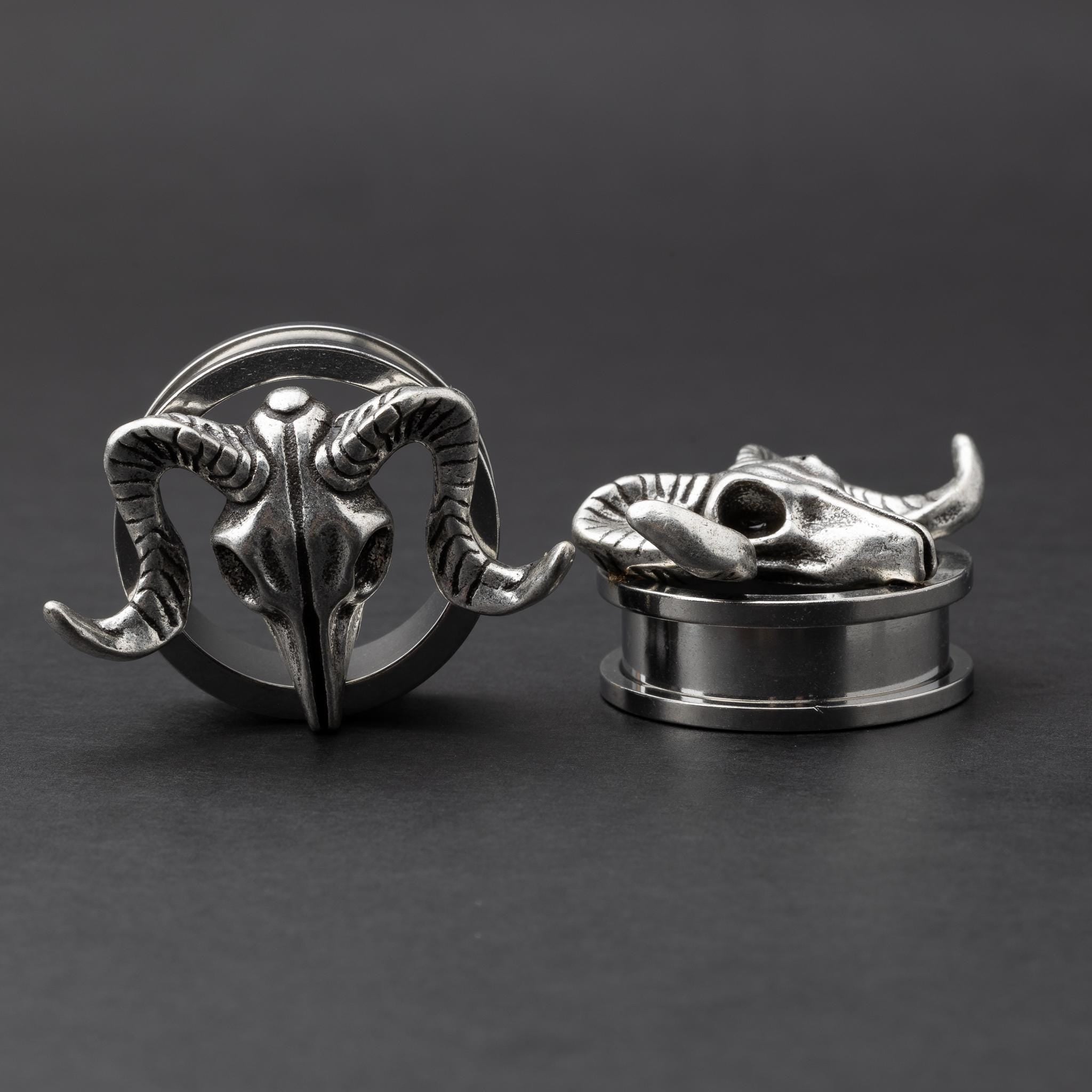 A pair of stainless steel saddle plugs featuring a 3D ram skull design, displayed on a dark, textured background.