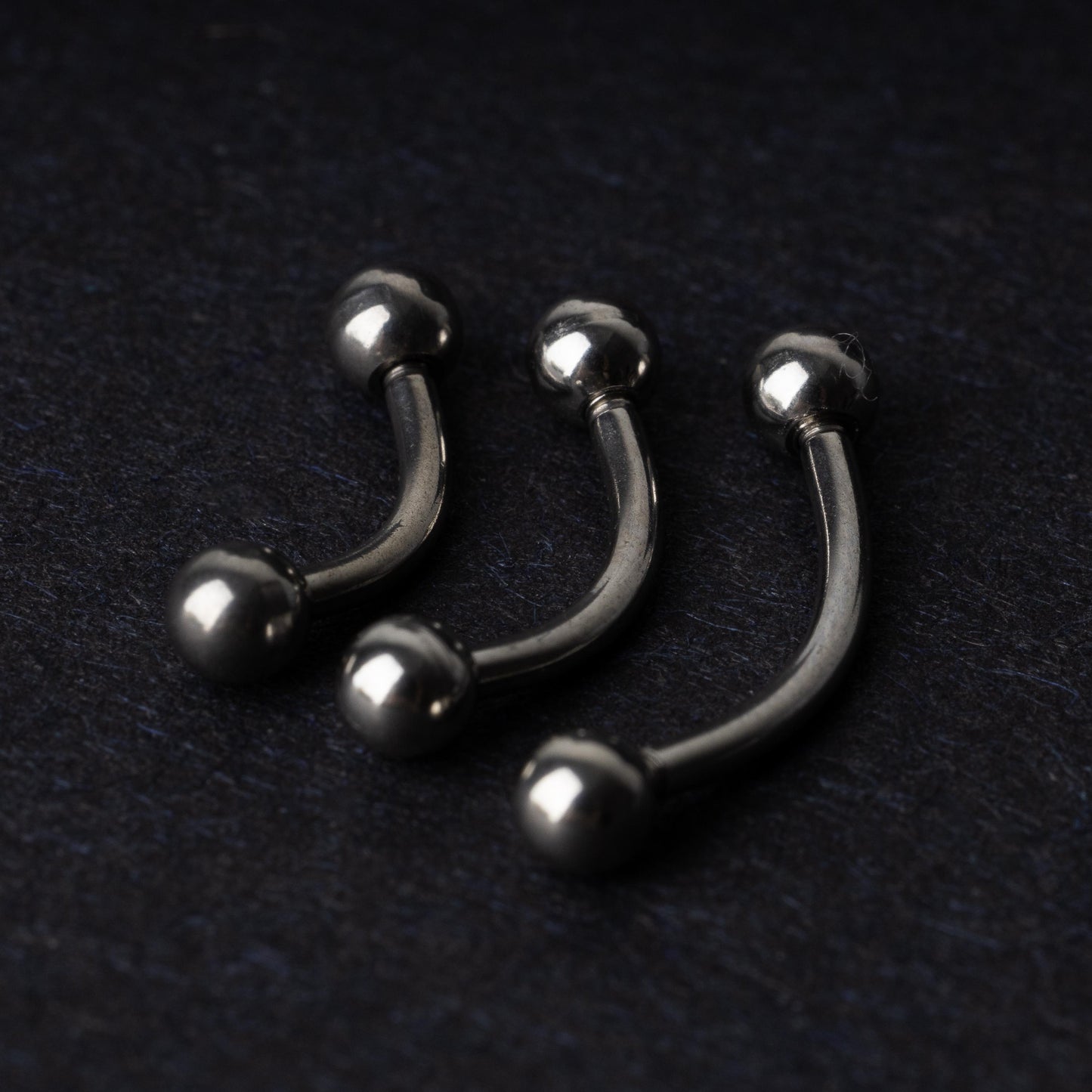 Three silver stainless steel curved barbells with polished ball ends, displayed on a dark textured background.
