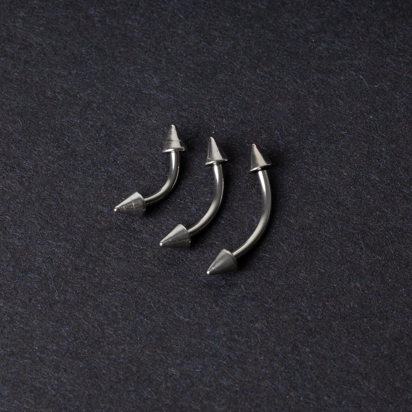 Three stainless steel curved barbells with cone-shaped ends, displayed on a dark background.