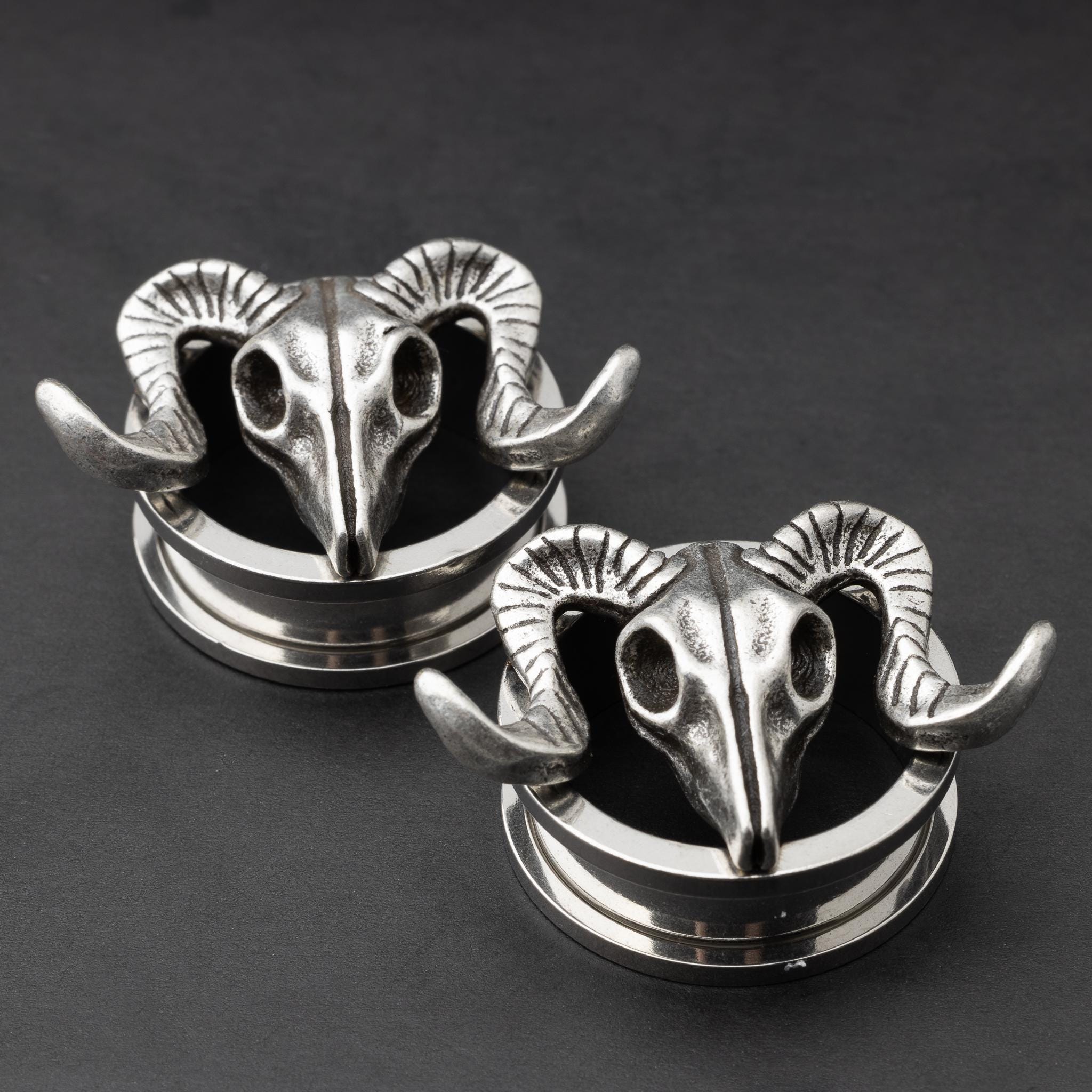 A pair of stainless steel saddle plugs featuring a 3D ram skull design, displayed on a dark, textured background.