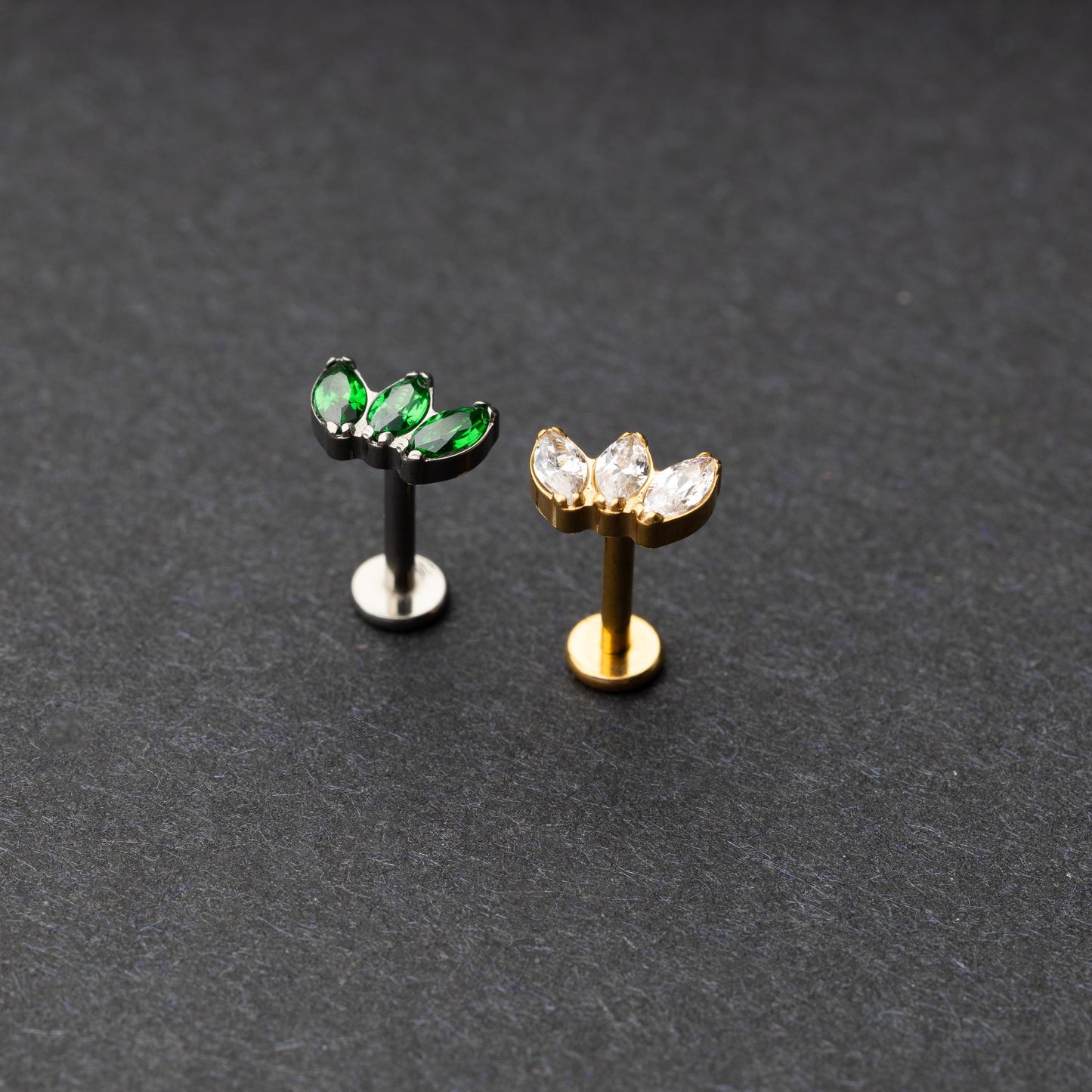 Two flat back bars featuring a triple gem design: one with emerald green gems on a silver base and the other with clear gems on a gold base, displayed on a dark background.