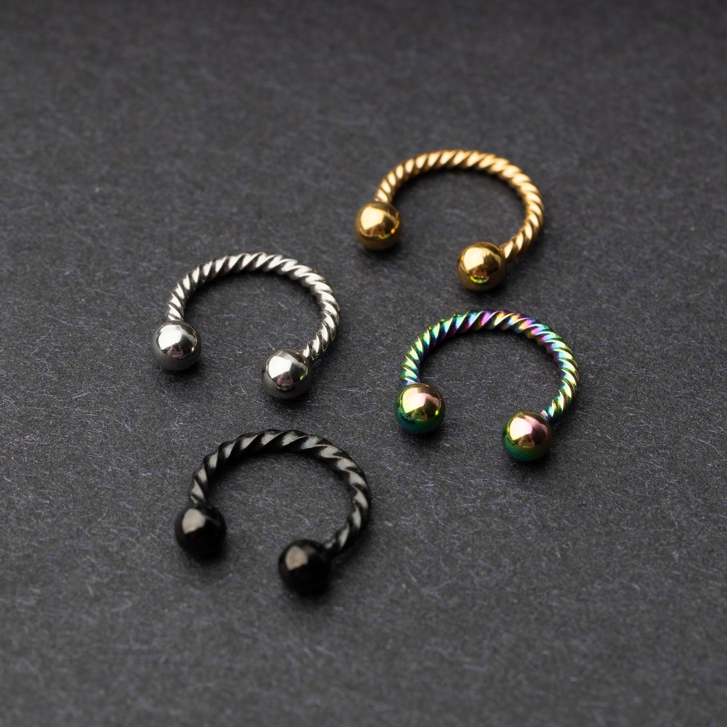 Twisted horseshoe barbells in silver, rainbow, gold, and black finishes displayed on a dark background, highlighting their bold and unique design.