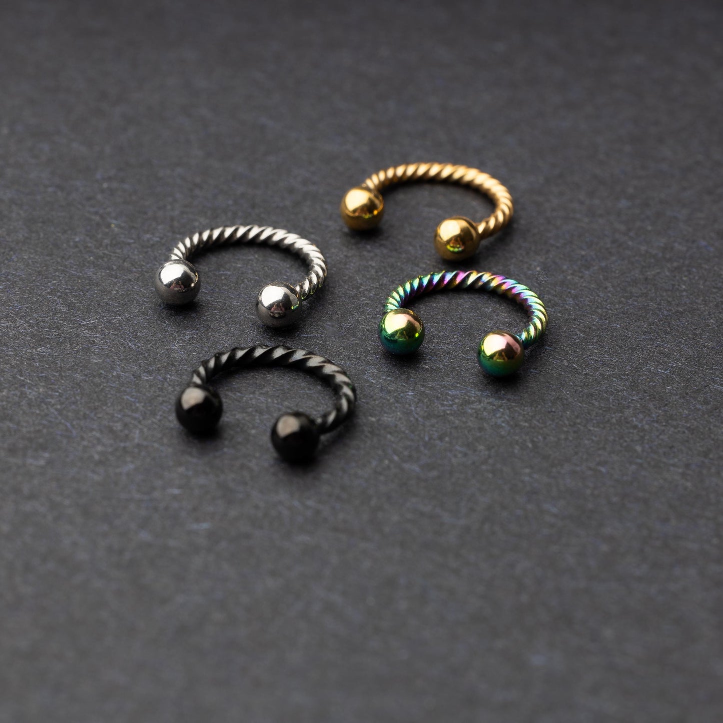 Twisted horseshoe barbells in silver, rainbow, gold, and black finishes displayed on a dark background, highlighting their bold and unique design.