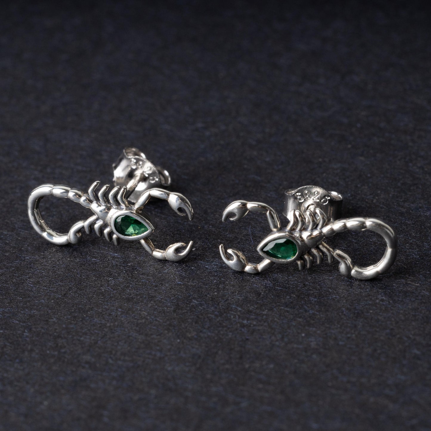 A pair of 925 sterling silver scorpion-shaped earrings with green gemstone accents as eyes, displayed on a dark background.