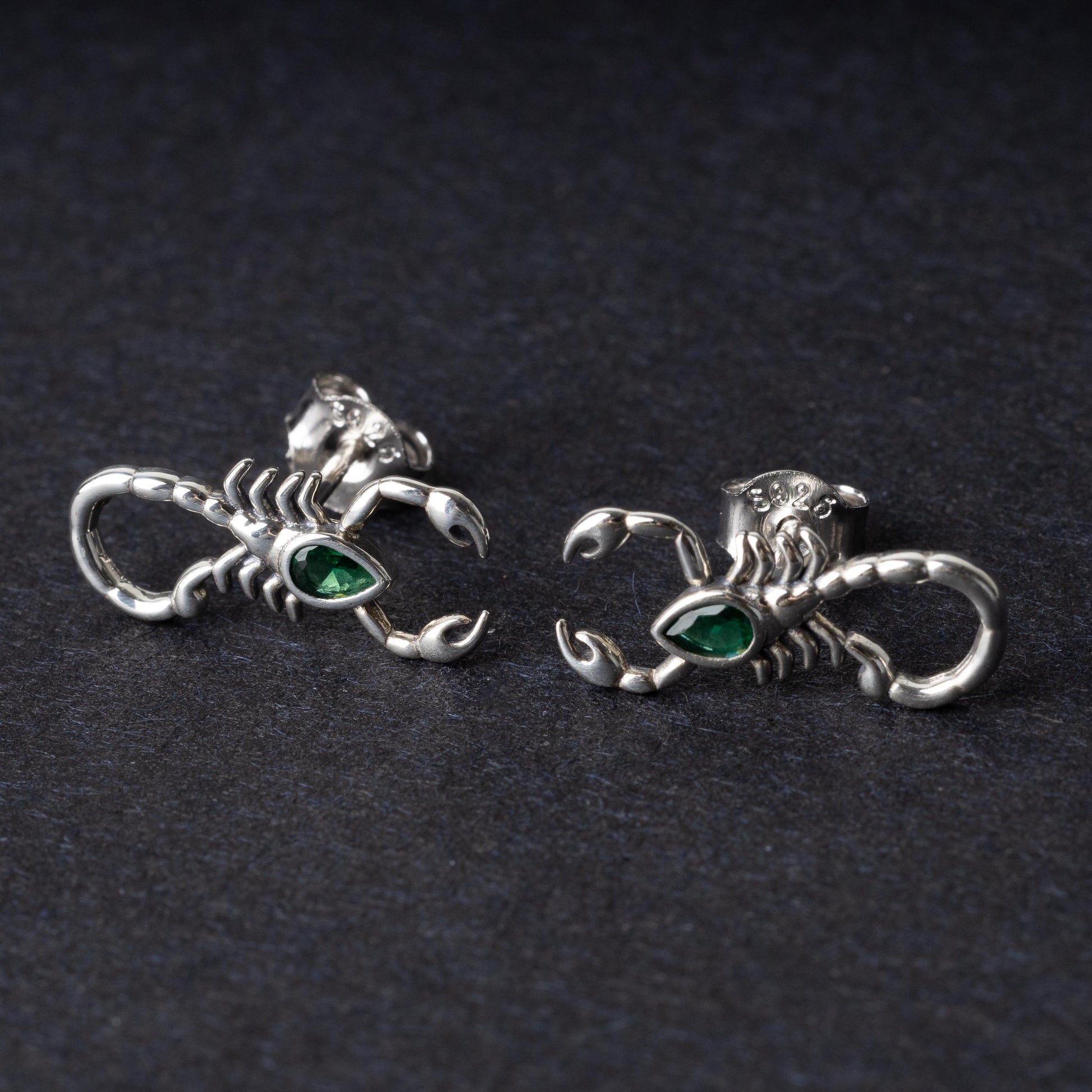 A pair of 925 sterling silver scorpion-shaped earrings with green gemstone accents as eyes, displayed on a dark background.
