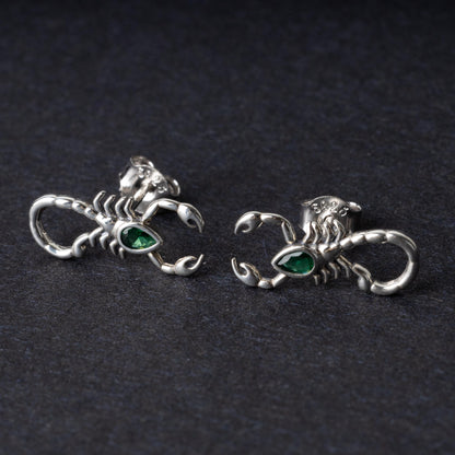 A pair of 925 sterling silver scorpion-shaped earrings with green gemstone accents as eyes, displayed on a dark background.
