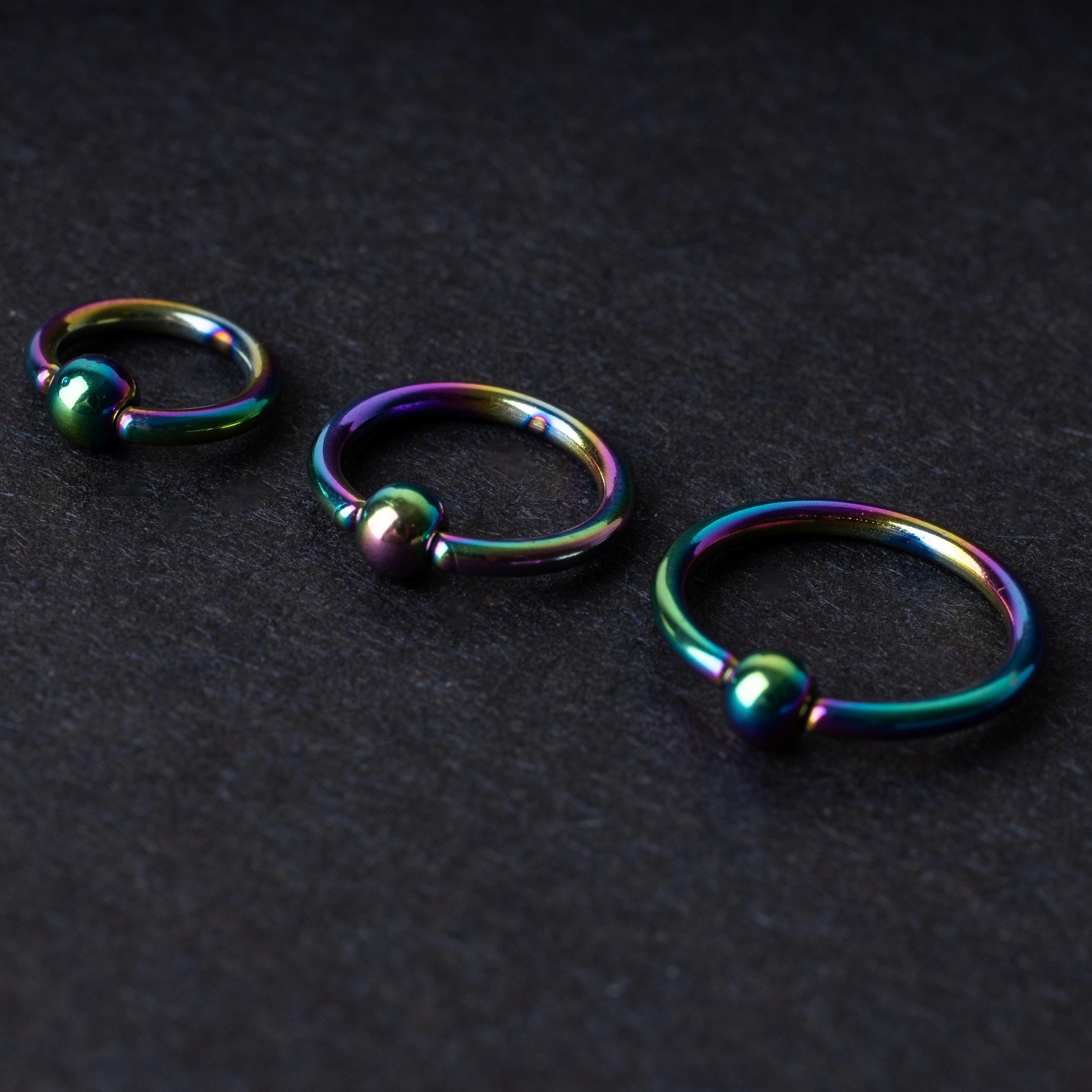 Three iridescent titanium captive bead rings of varying sizes displayed on a dark textured background, showcasing their vibrant rainbow finish.