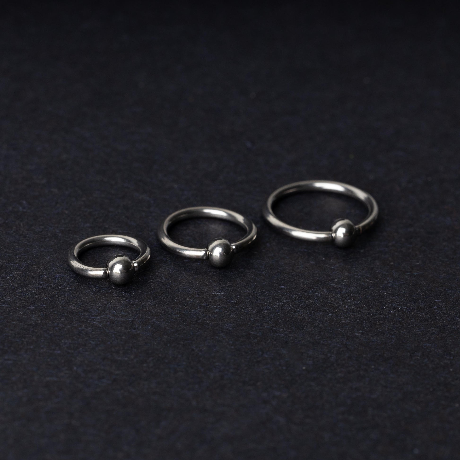 Three polished stainless steel captive bead rings of varying sizes displayed on a dark matte background.
