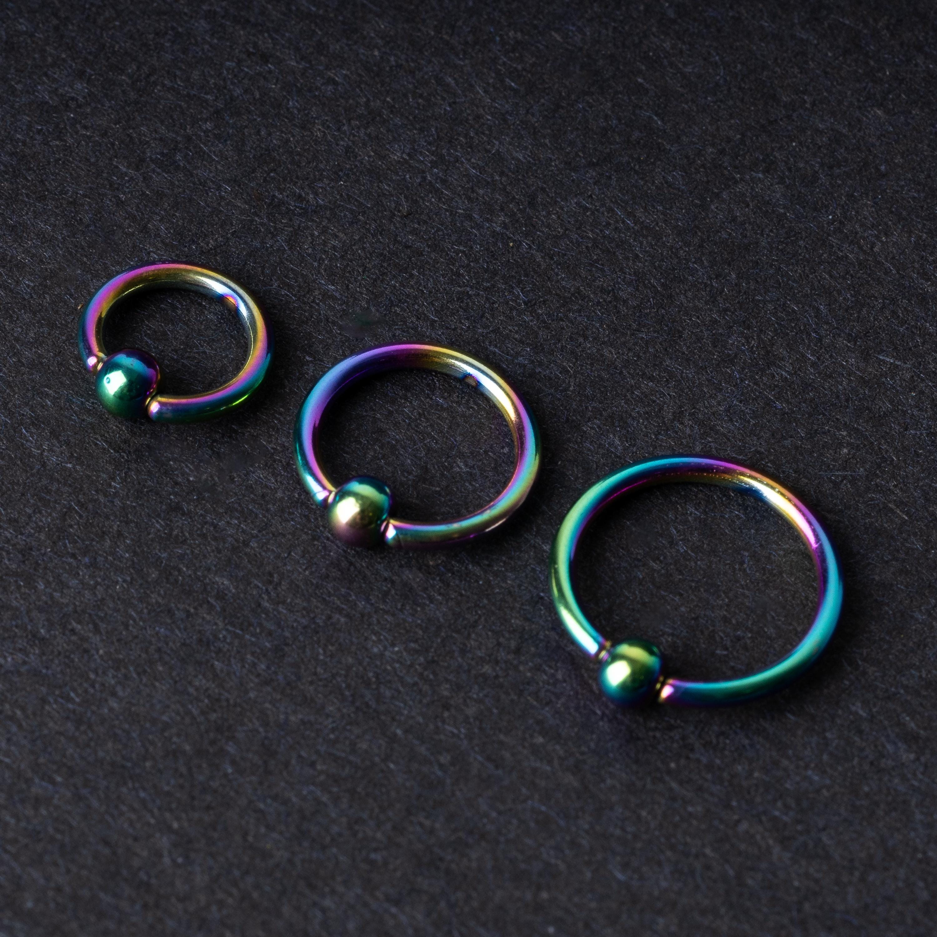 Three iridescent titanium captive bead rings of varying sizes displayed on a dark textured background, showcasing their vibrant rainbow finish.