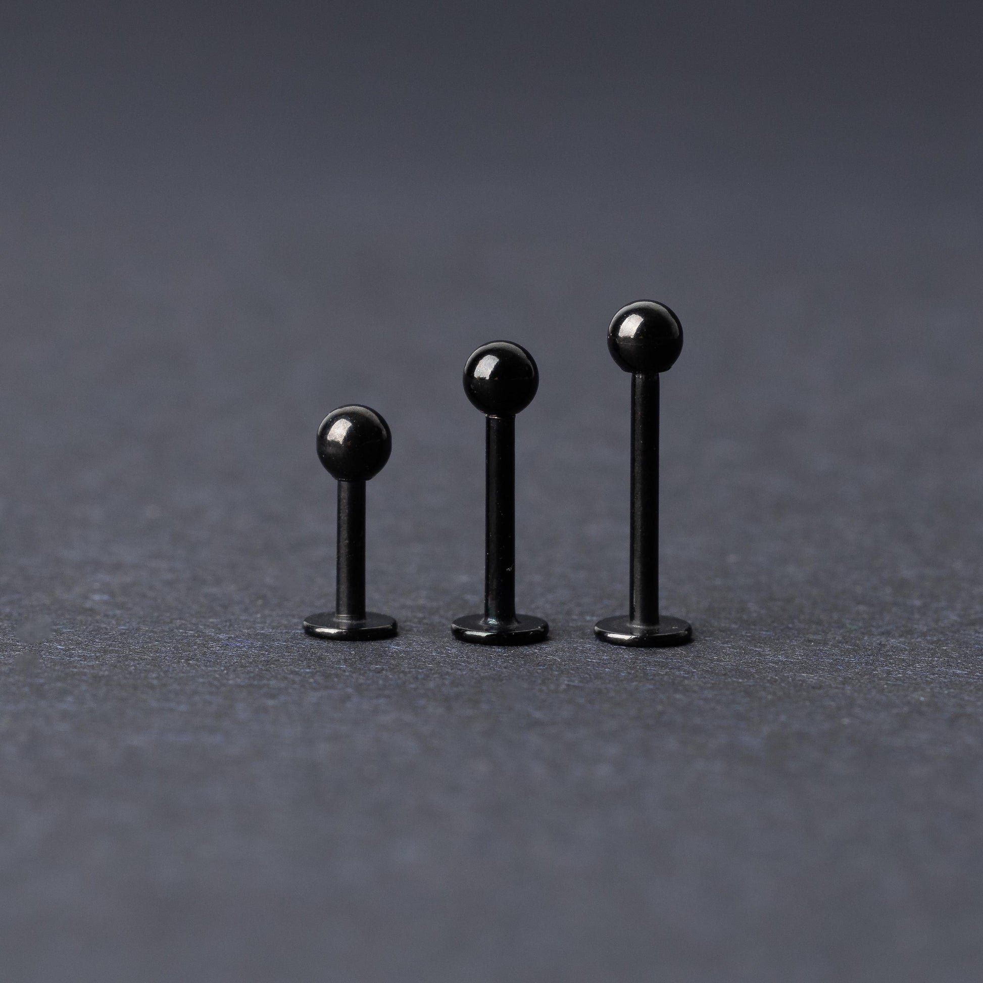 Three black stainless steel flat back barbells with polished finishes, displayed on a dark background.