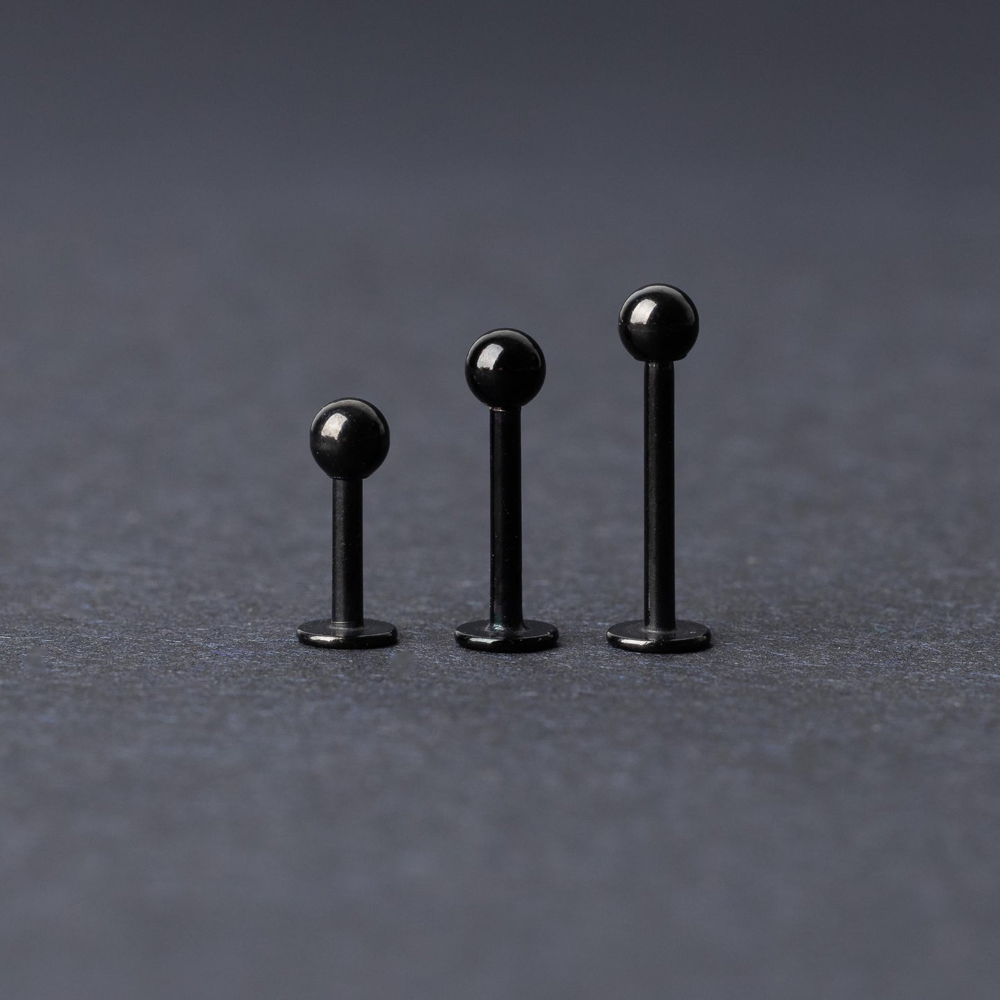 Three black stainless steel flat back barbells with polished finishes, displayed on a dark background.
