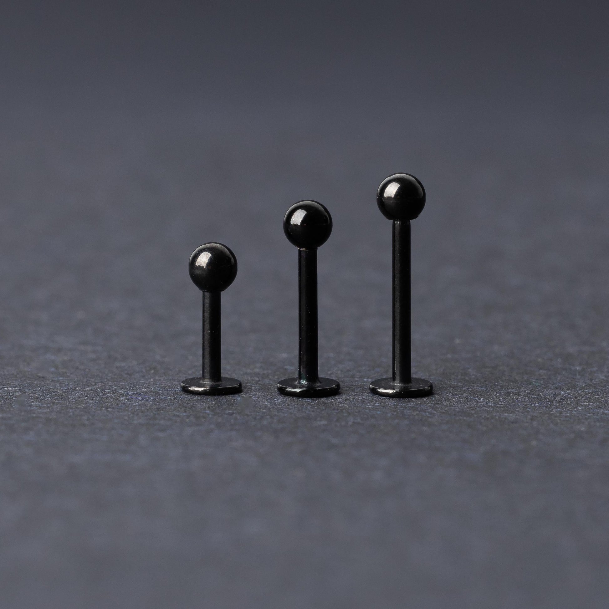 Three black stainless steel flat back barbells with polished finishes, displayed on a dark background.