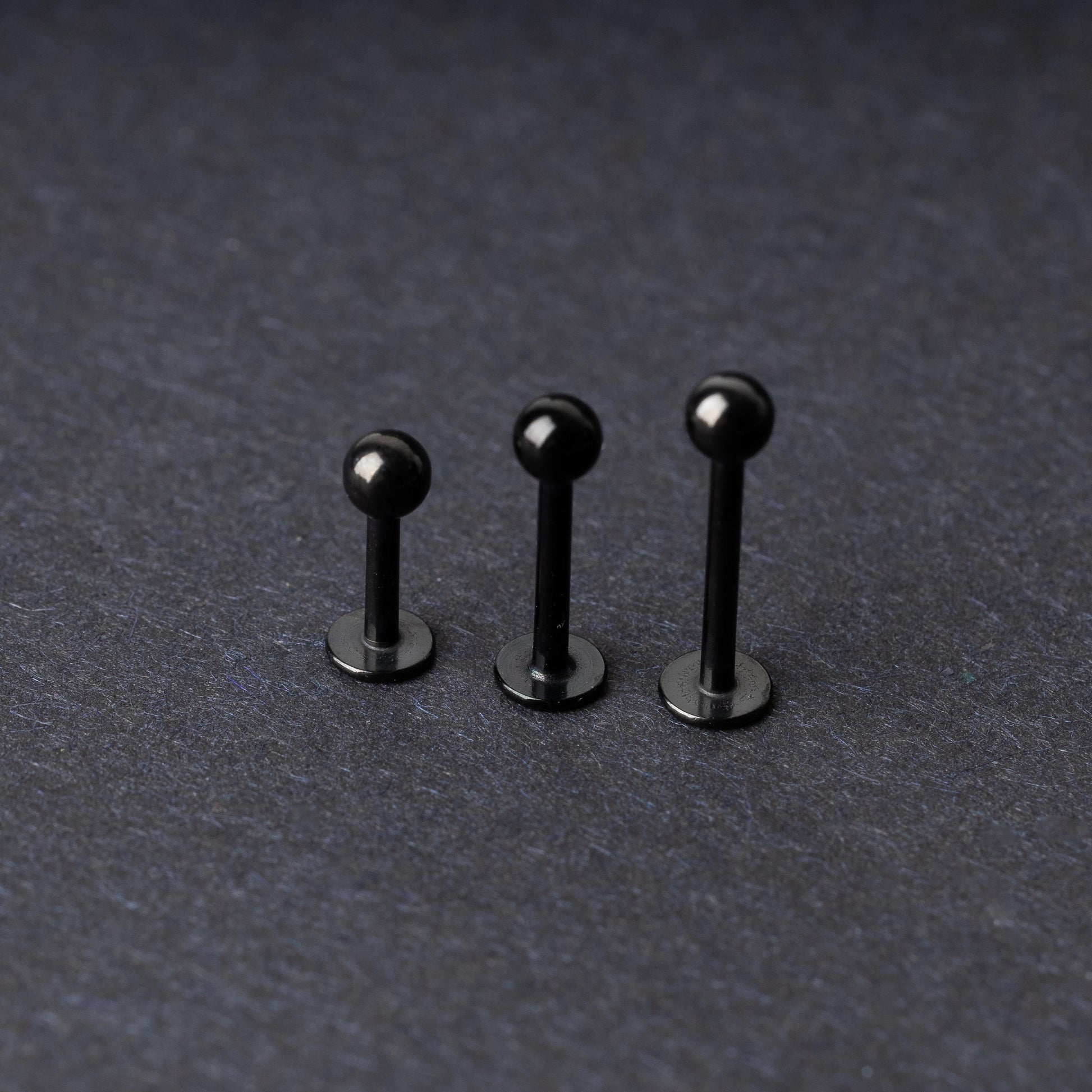 Three black stainless steel flat back barbells with polished finishes, displayed on a dark background.