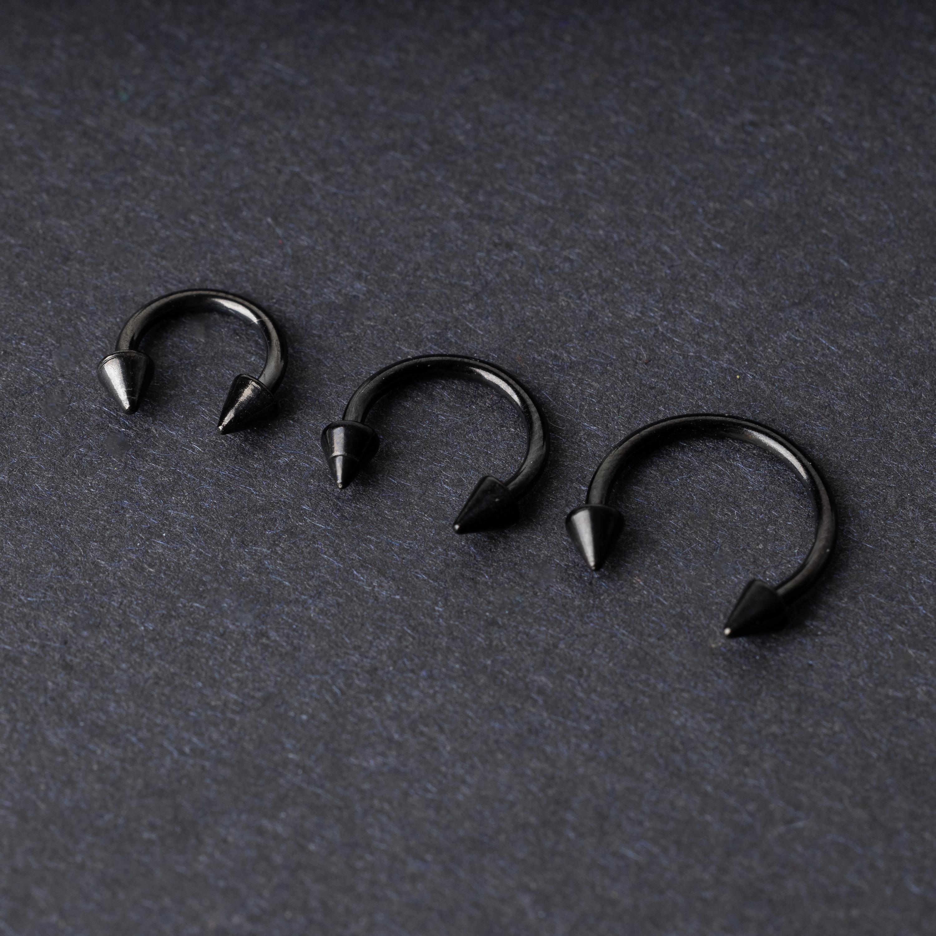 Three black stainless steel horseshoe rings with spiked ends in varying sizes, displayed on a dark background.