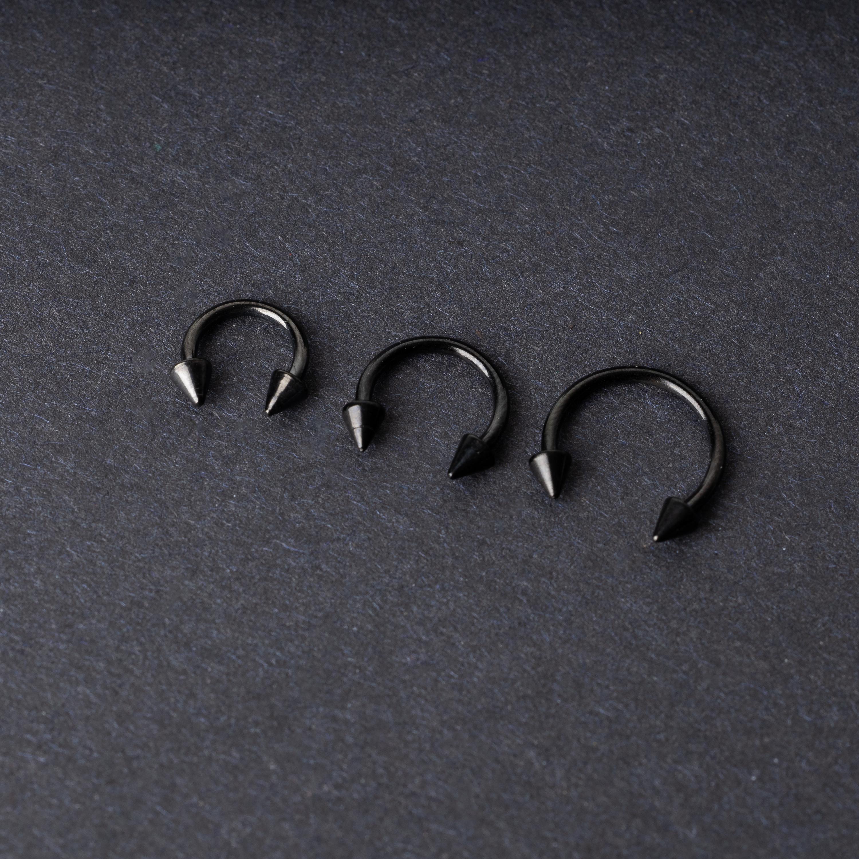 Three black stainless steel horseshoe rings with spiked ends in varying sizes, displayed on a dark background.