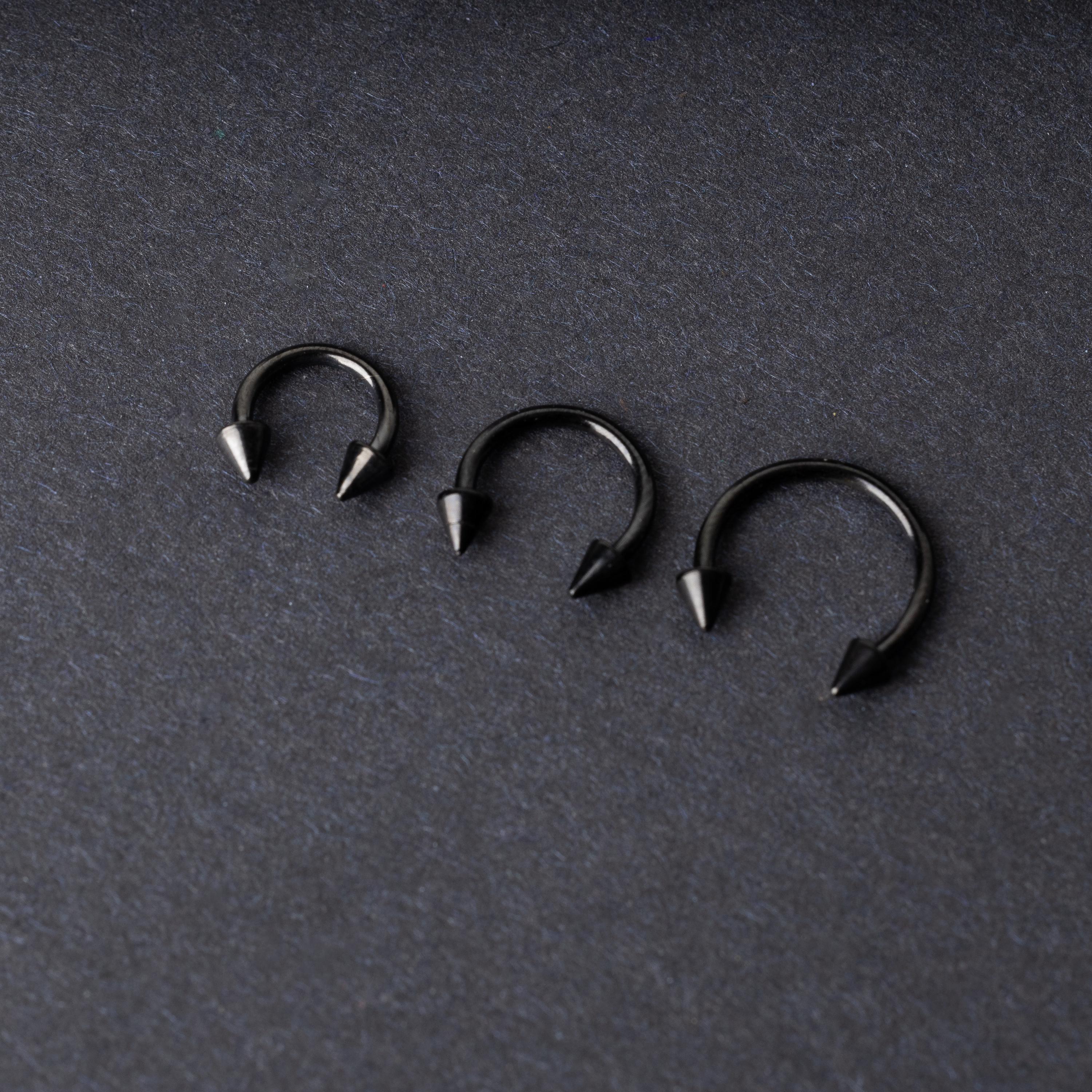 Three black stainless steel horseshoe rings with spiked ends in varying sizes, displayed on a dark background.