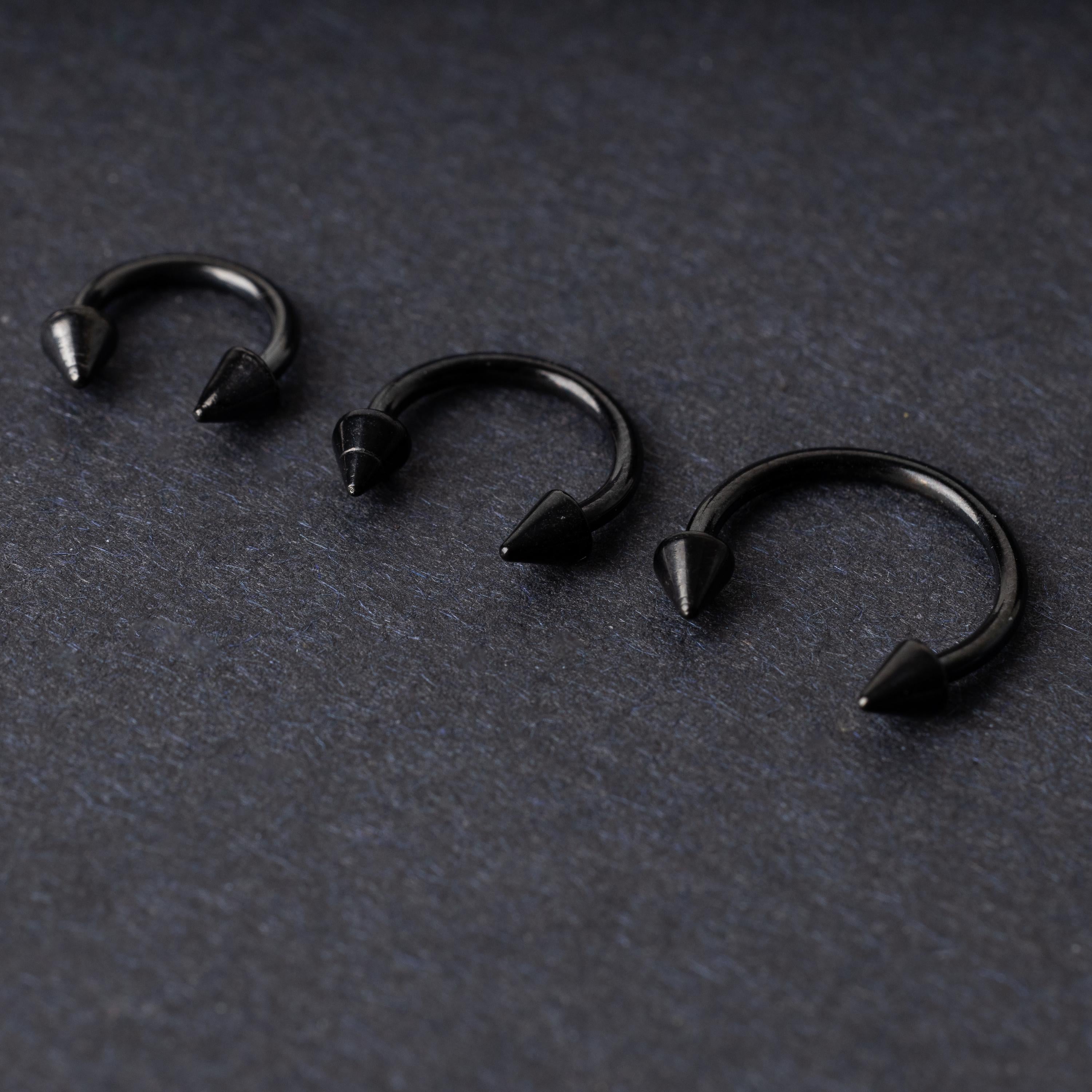 Three black stainless steel horseshoe rings with spiked ends in varying sizes, displayed on a dark background.