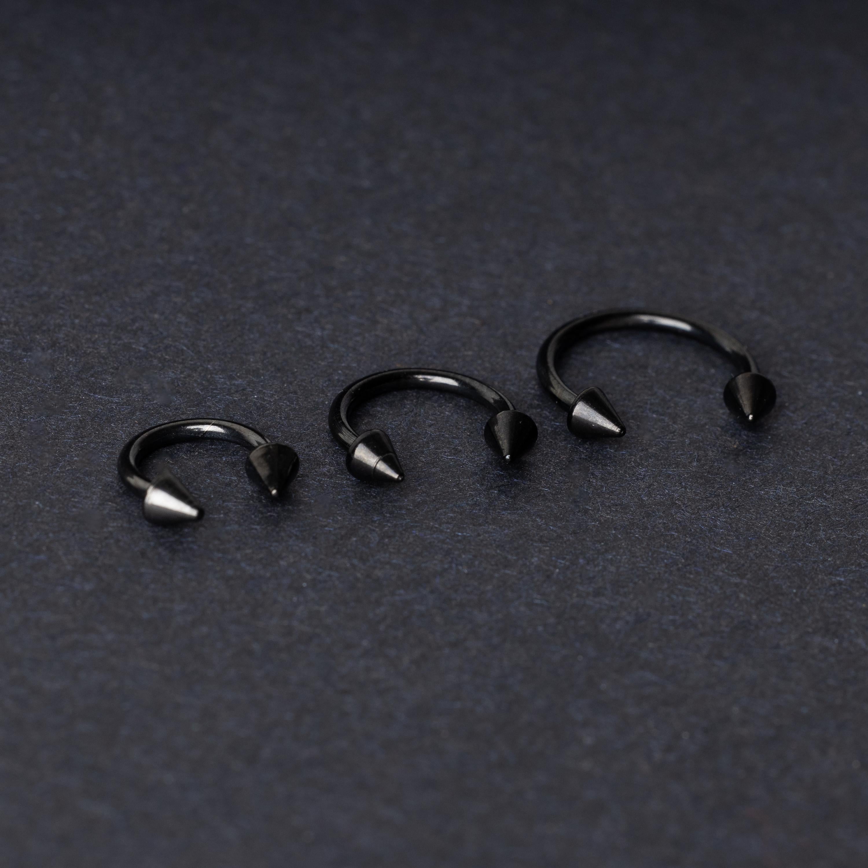 Three black stainless steel horseshoe rings with spiked ends in varying sizes, displayed on a dark background.