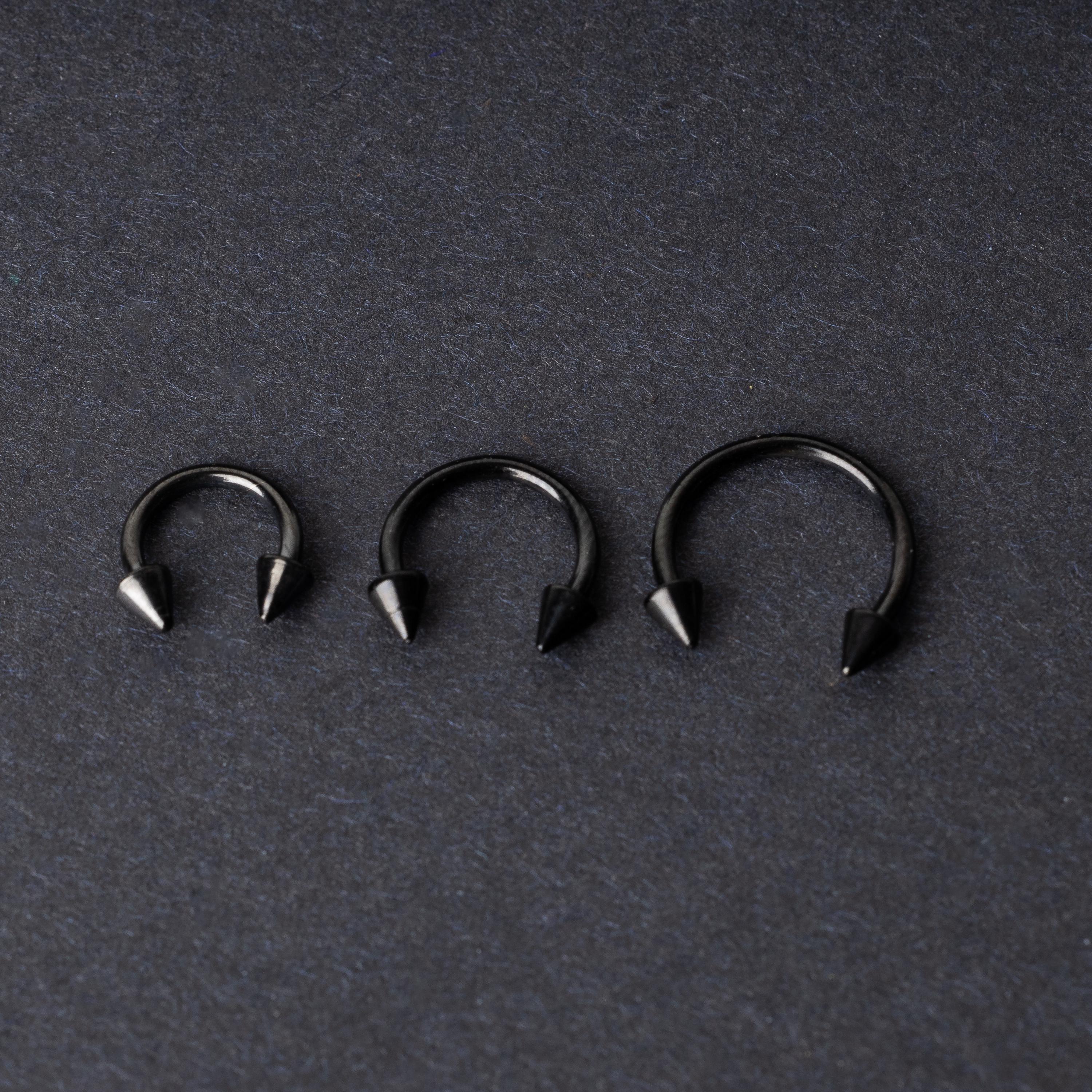 Three black stainless steel horseshoe rings with spiked ends in varying sizes, displayed on a dark background.