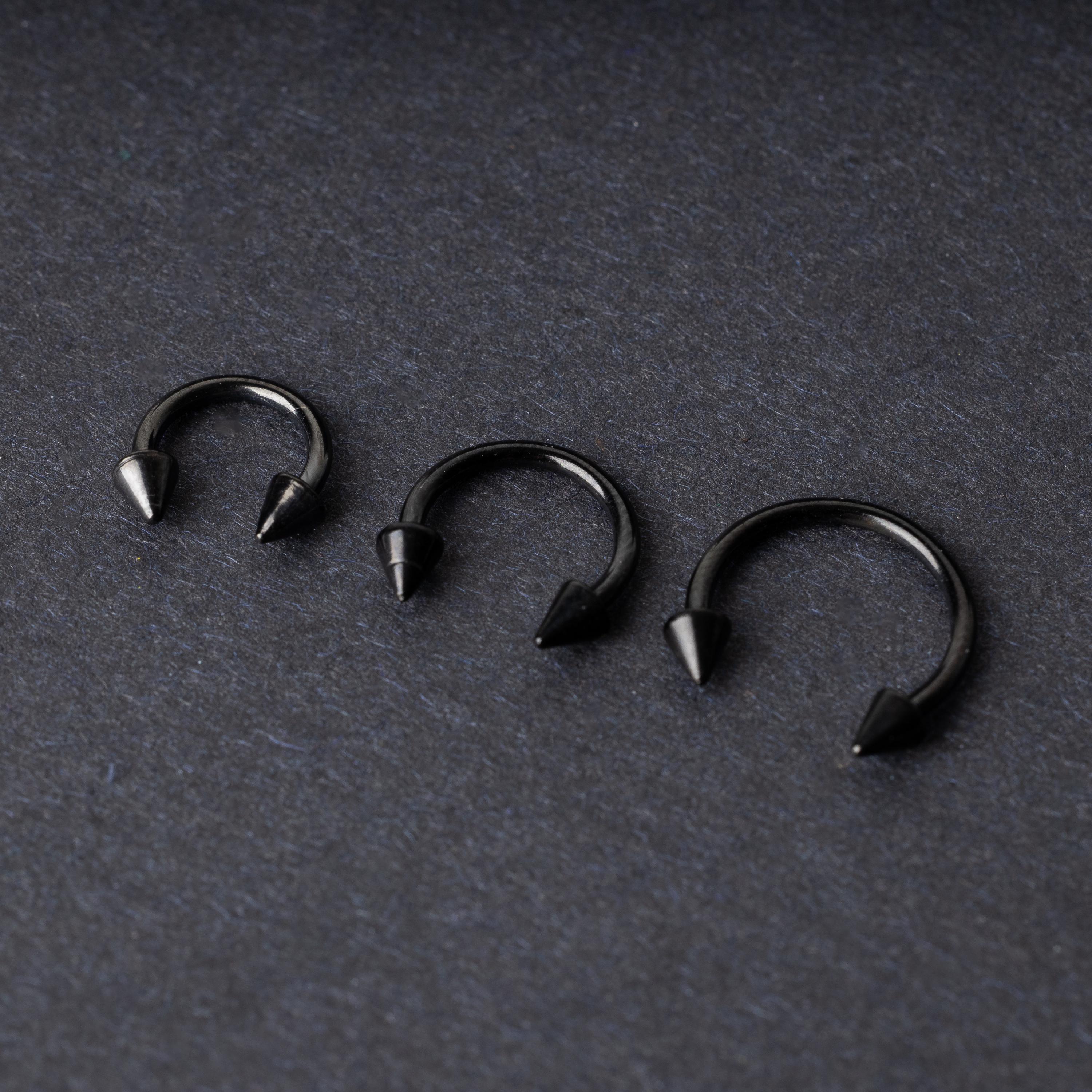 Three black stainless steel horseshoe rings with spiked ends in varying sizes, displayed on a dark background.
