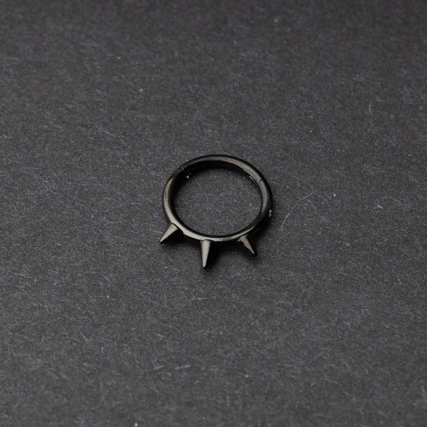 Black-tone stainless steel clicker ring with three spikes, displayed on a dark textured background.