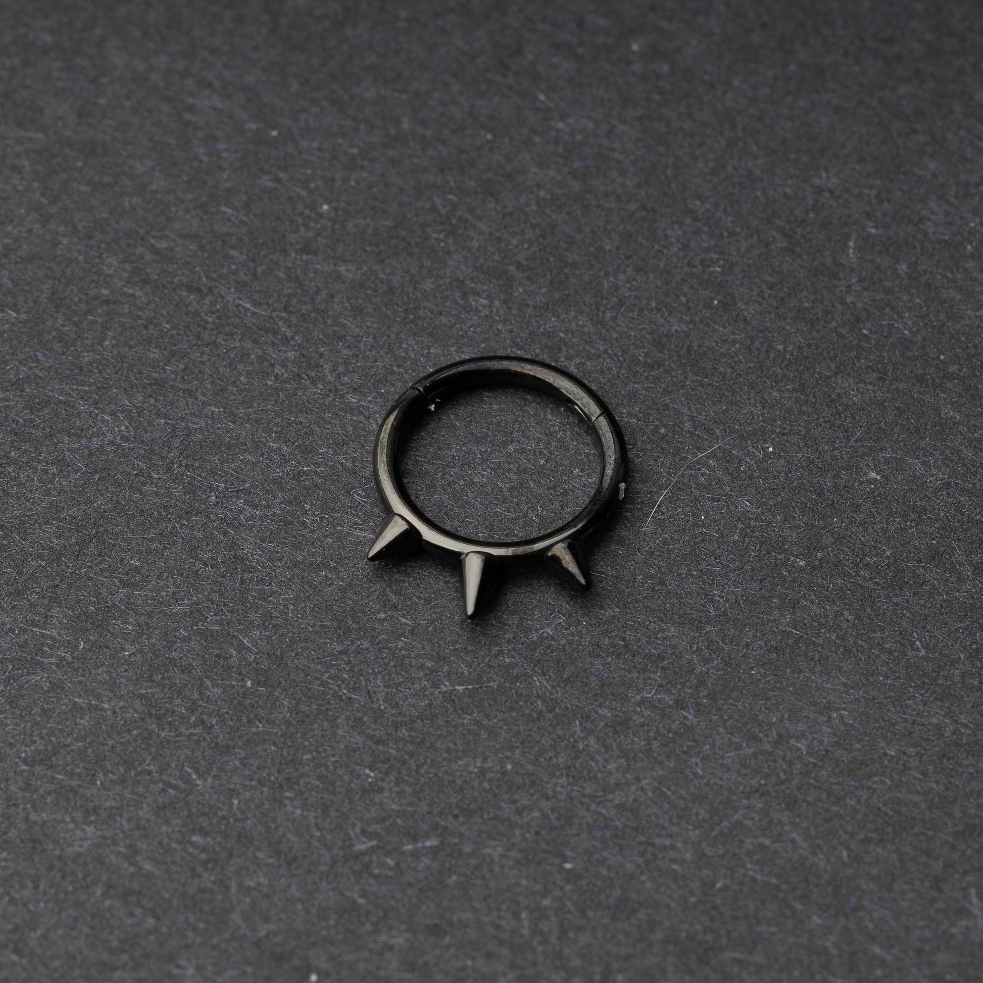 Black-tone stainless steel clicker ring with three spikes, displayed on a dark textured background.