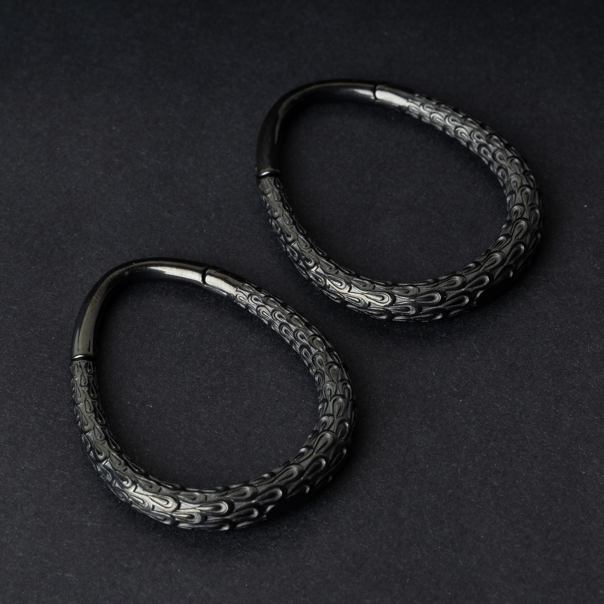 A pair of black teardrop ear weights featuring a detailed dragon-scale texture and a seamless hinge closure, displayed on a dark background.
