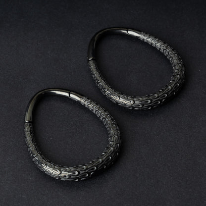 A pair of black teardrop ear weights featuring a detailed dragon-scale texture and a seamless hinge closure, displayed on a dark background.
