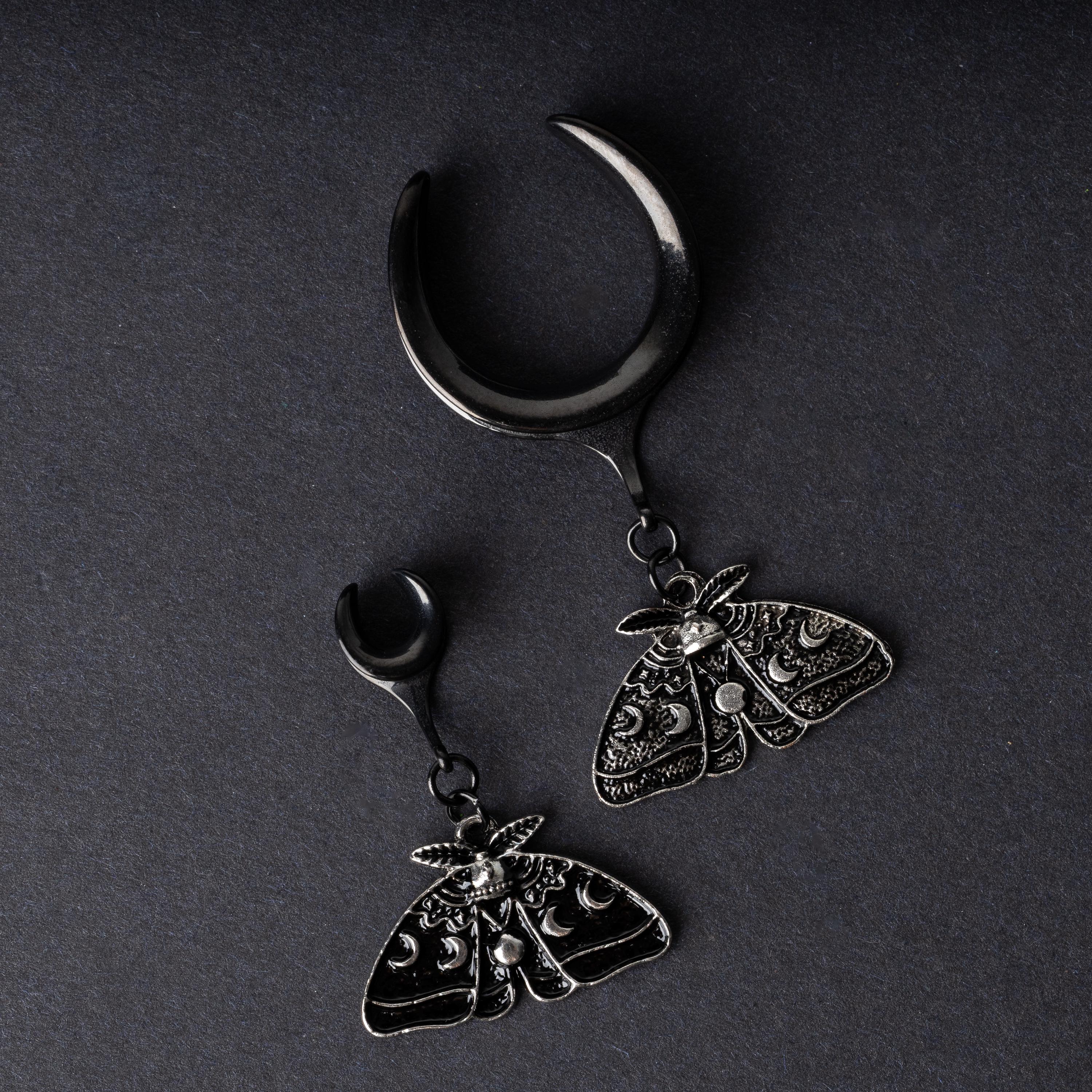 Black tunnels for stretched ears in two different sizes with dangling ornate moth charms featuring celestial patterns, displayed on a dark background.